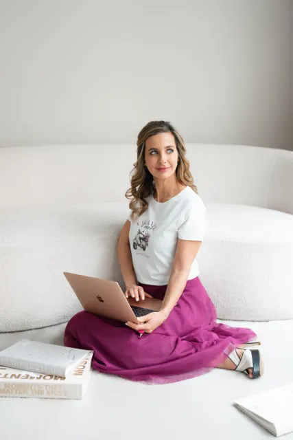 Frau sitzt auf weißem Boden vor Sofa, nutzt Laptop auf Schoß, trägt weißes T-Shirt und langen lila Rock, Blick nach rechts gerichtet, Bücher liegen in der Nähe.
