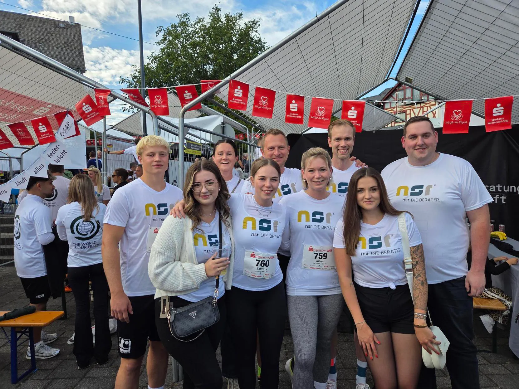 Group of eight adults wearing matching white NSR Die Berater team shirts posing outdoors under a tent with red Sparkasse Westerwald-Sieg banners above them.