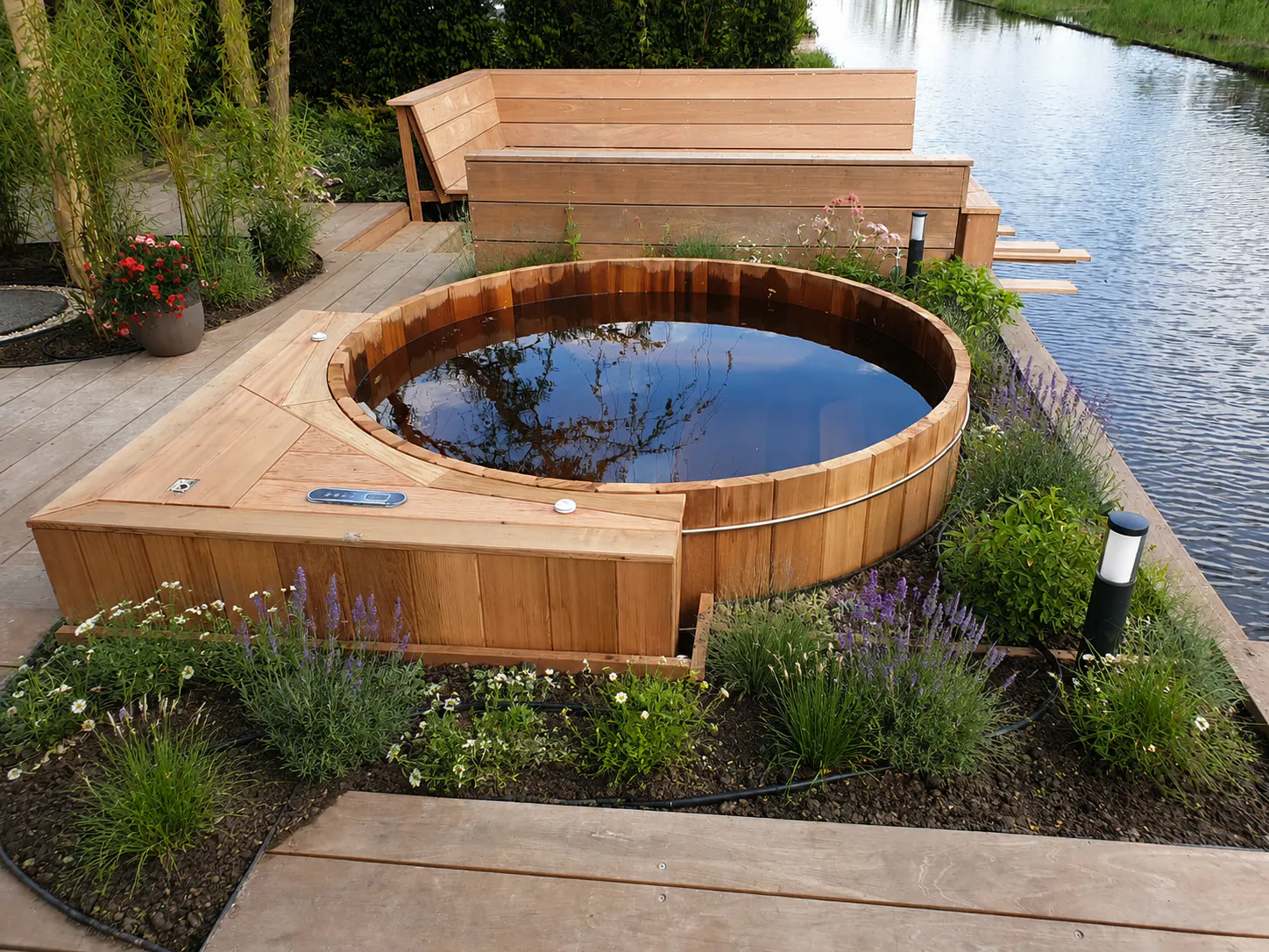 Wooden hot tub filled with water reflecting the sky, surrounded by plants and flowers, next to a lake and wooden outdoor seating area.