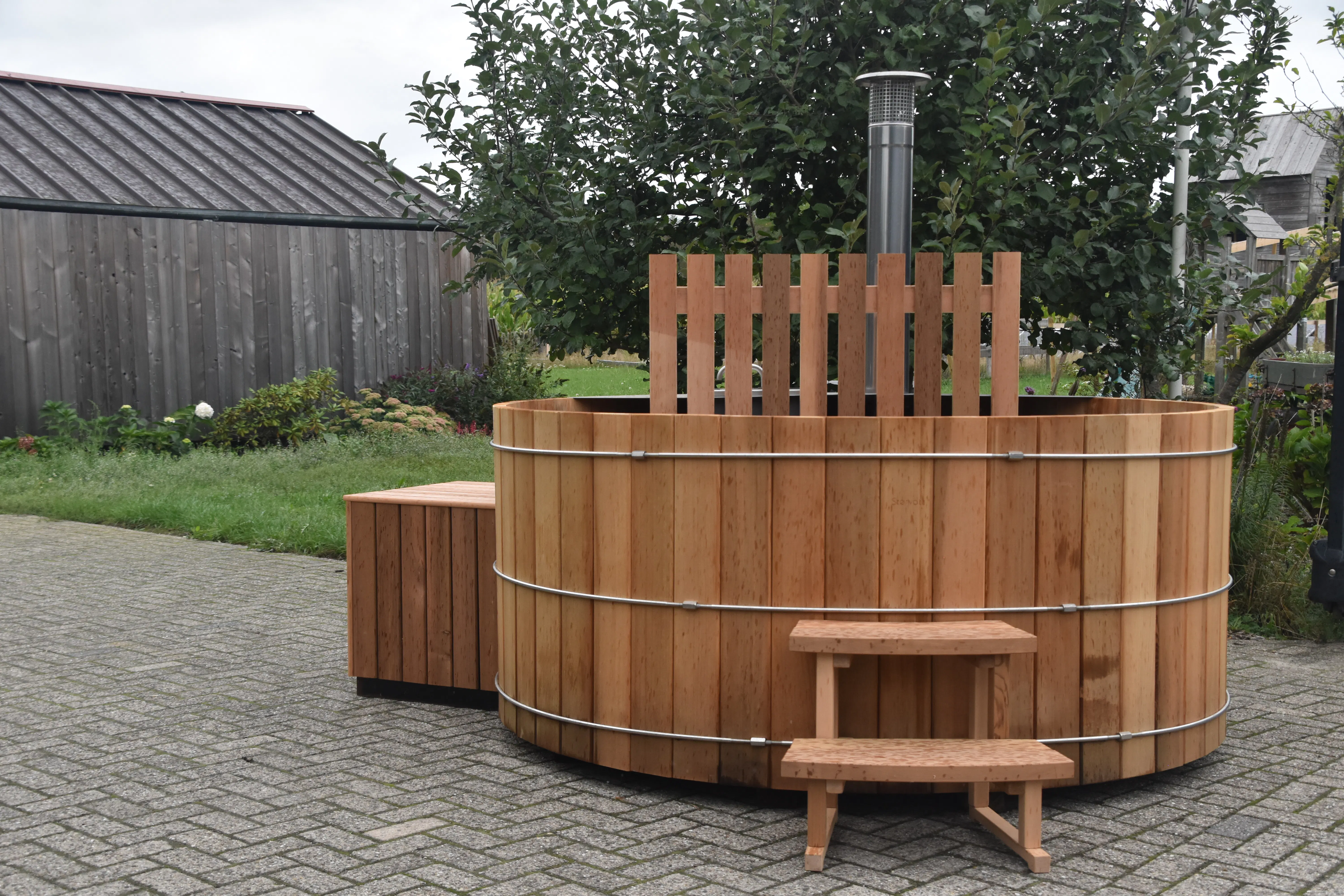 Circular wooden outdoor hot tub with metal bands and small wooden steps, placed on paved ground near a wooden shed and garden greenery.
