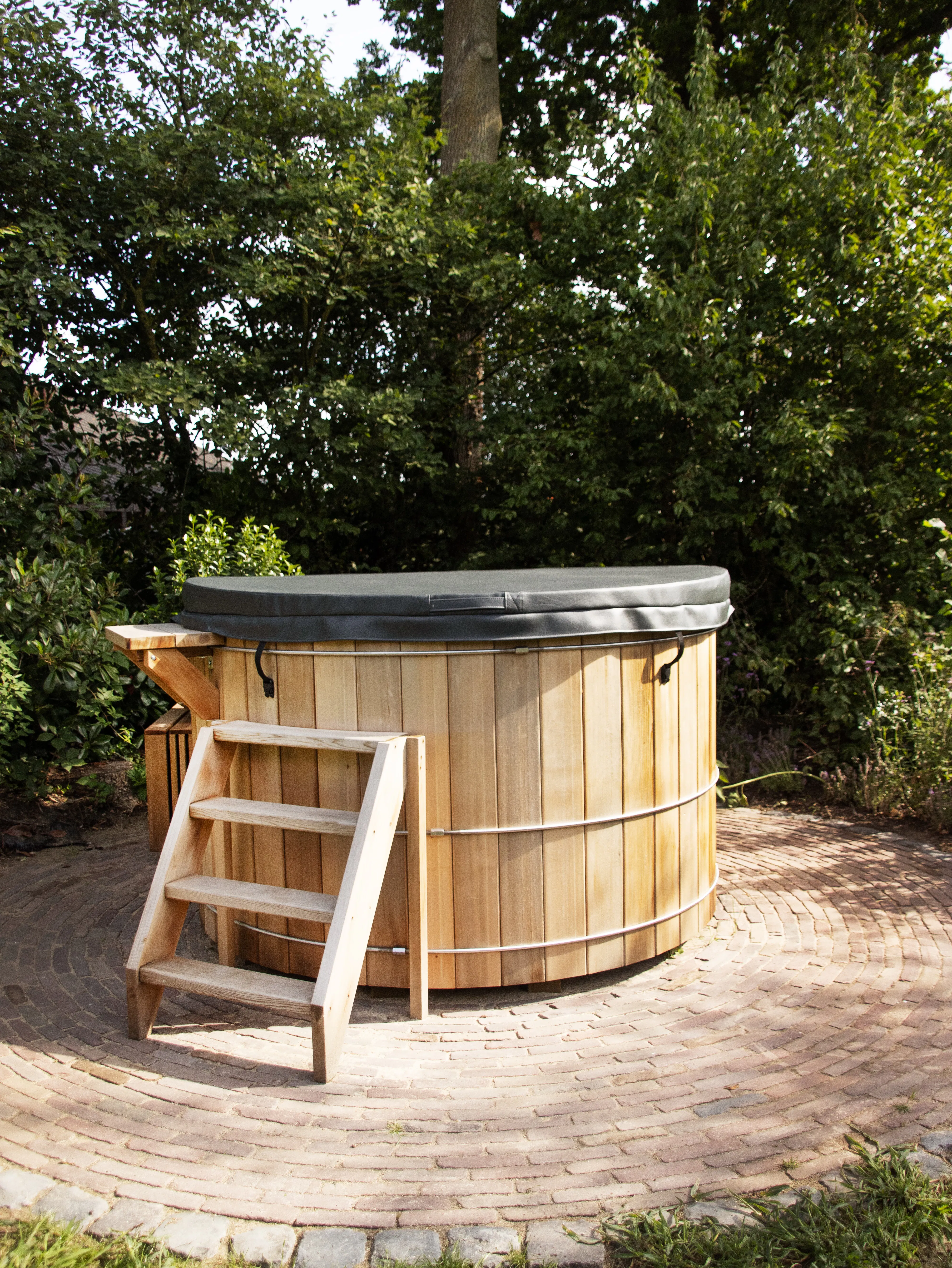 Circular wooden hot tub with a gray cover and wooden steps, placed on a circular brick patio surrounded by green bushes and trees.