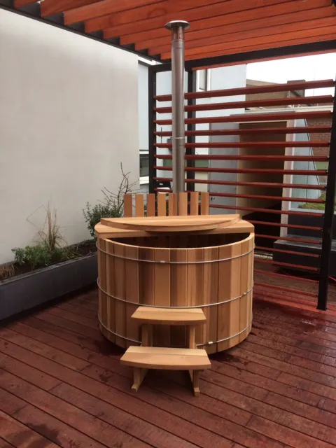 Wooden round hot tub with metal chimney pipe on a wooden deck, next to a planter with small plants, under a wooden pergola.