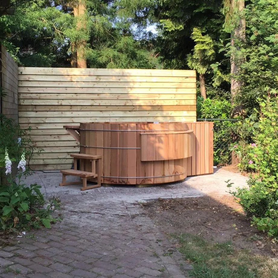 Wooden outdoor hot tub with small steps in a garden area, surrounded by plants, trees, and a wooden privacy fence.