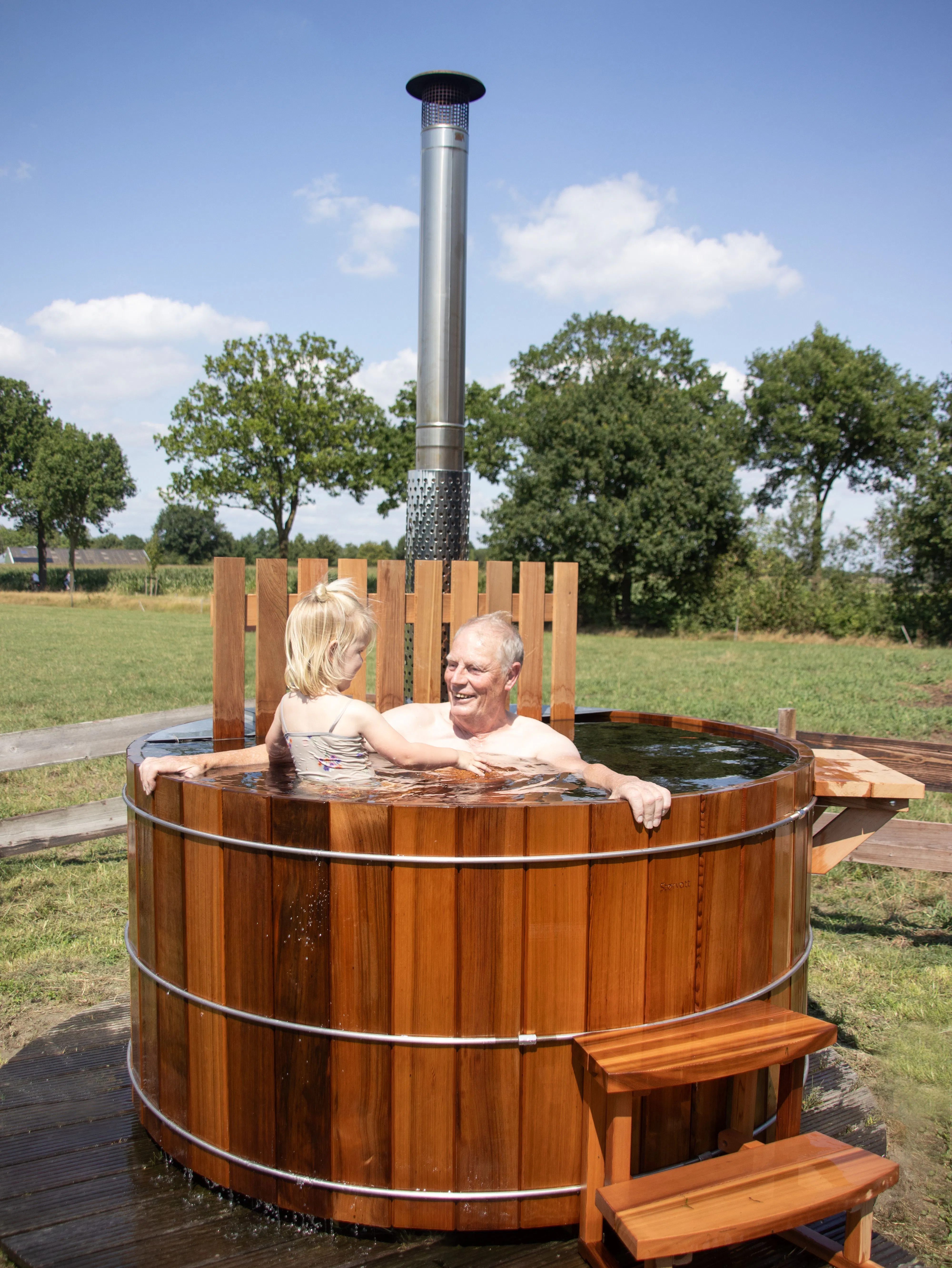 Elderly man and young child sitting in a round wooden hot tub with a metal chimney, outdoors on grass with trees and a blue sky in the background.