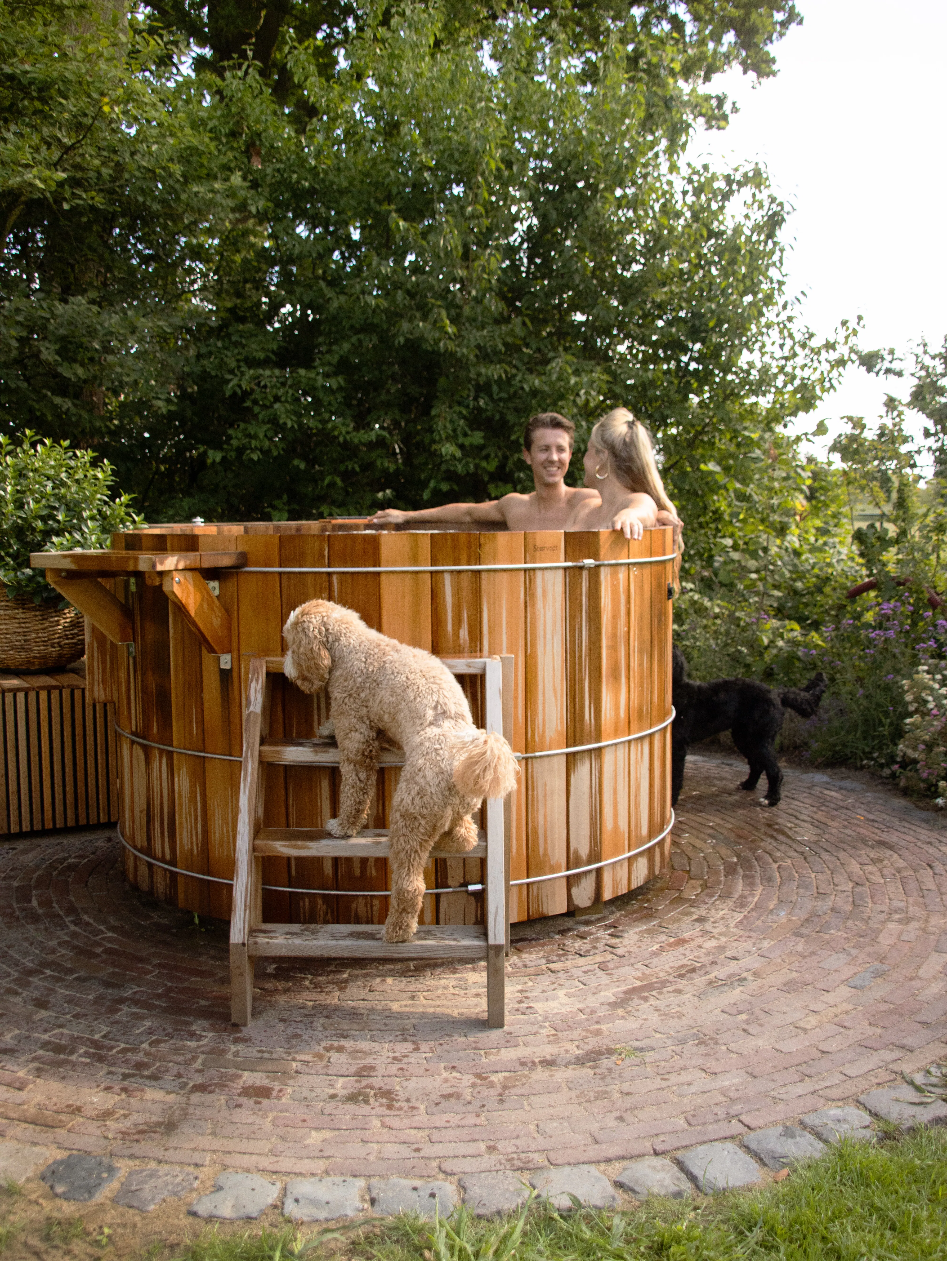 Two people sitting in a wooden outdoor hot tub with a curly dog climbing steps and another dog nearby, surrounded by trees and garden plants.