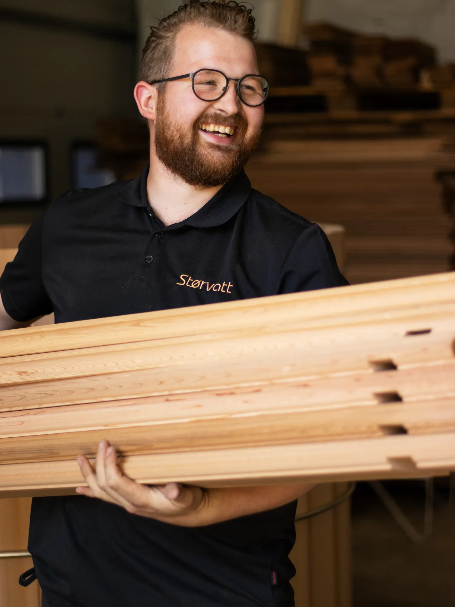Man in glasses and black polo shirt smiling while carrying stacked cedar planks in a workshop.
