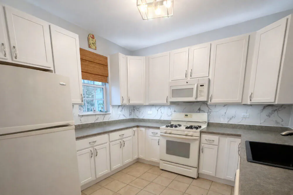 Kitchen with white cabinets, gray countertops, a white stove with overhead microwave, a white refrigerator, a black sink, and a window with a bamboo blind.