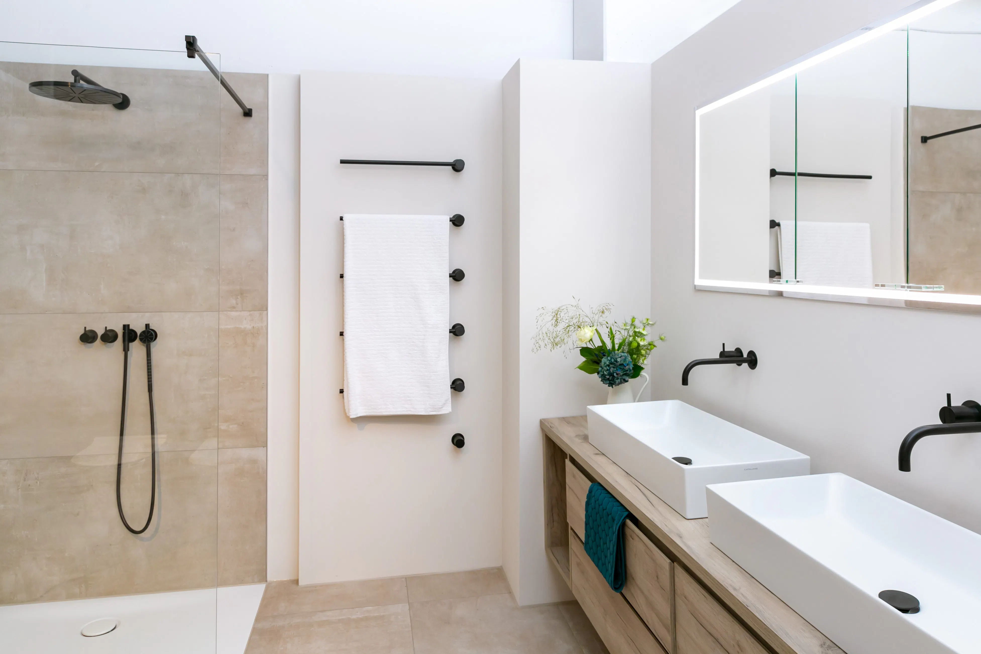 Moderner Badezimmerbereich mit begehbarer Dusche, zwei weißen rechteckigen Waschbecken auf Holzunterschrank, beleuchtetem Spiegel und weißem Handtuch an schwarzem Handtuchhalter.
