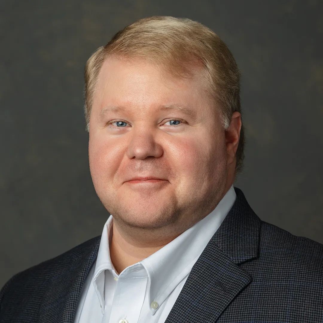 Close-up of a middle-aged man with light skin, blue eyes, short blonde hair, wearing a dark plaid blazer over a white button-up shirt, against a plain dark background.