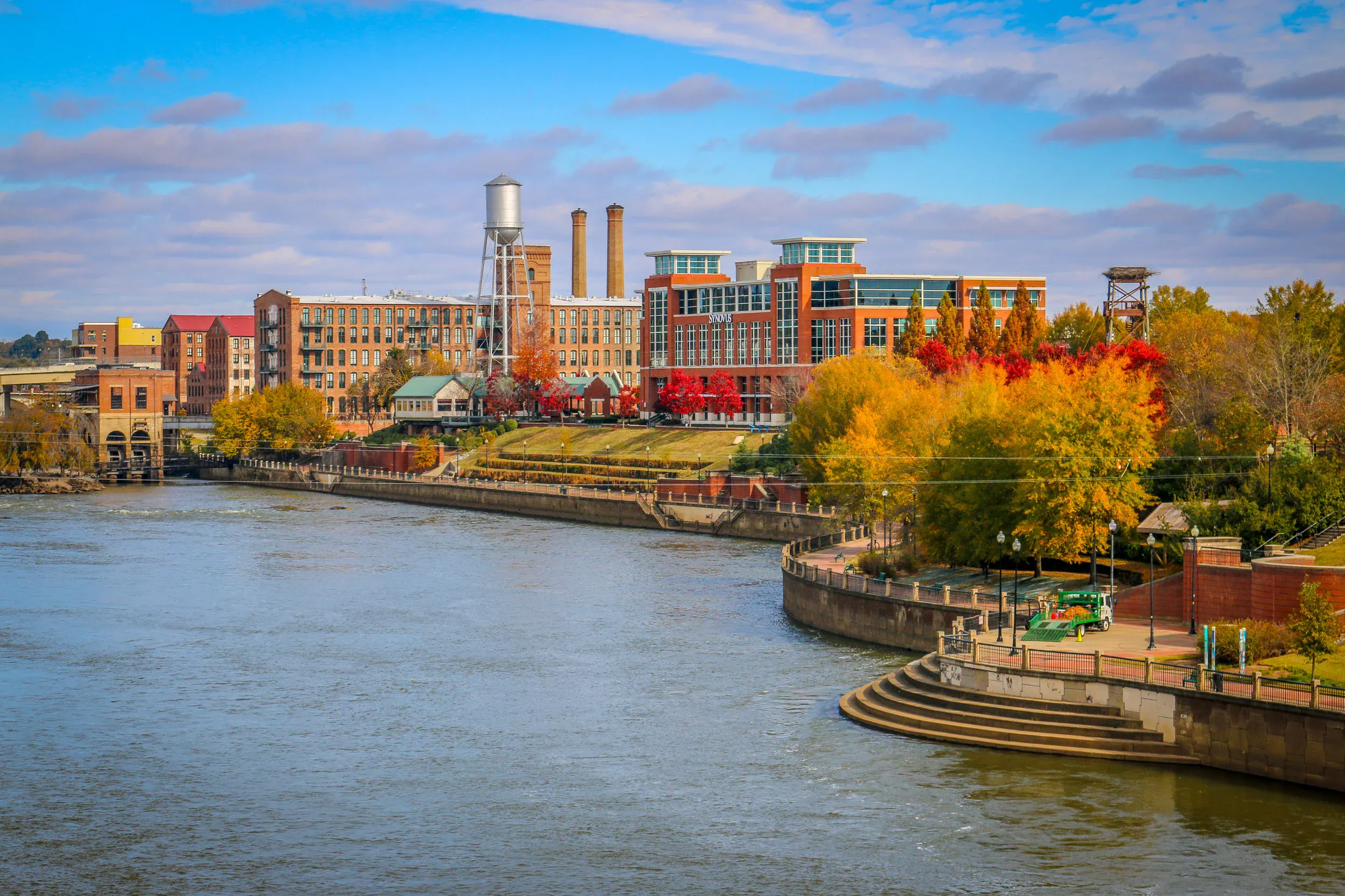 Riverside park with autumn trees, a curved concrete embankment, and brick buildings including a water tower and factory chimneys under a partly cloudy sky.