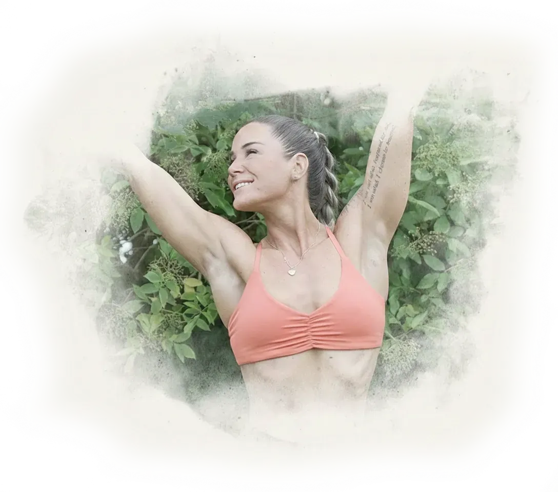 Woman in a coral sports bra with braided hair, smiling and raising her arms, standing in front of green leafy plants.