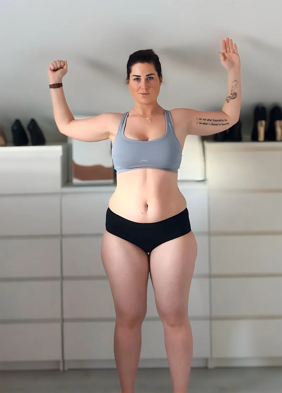 Woman standing in underwear and sports bra with arms raised, showing tattoos on her left arm, in a room with dressers and shoes in the background.