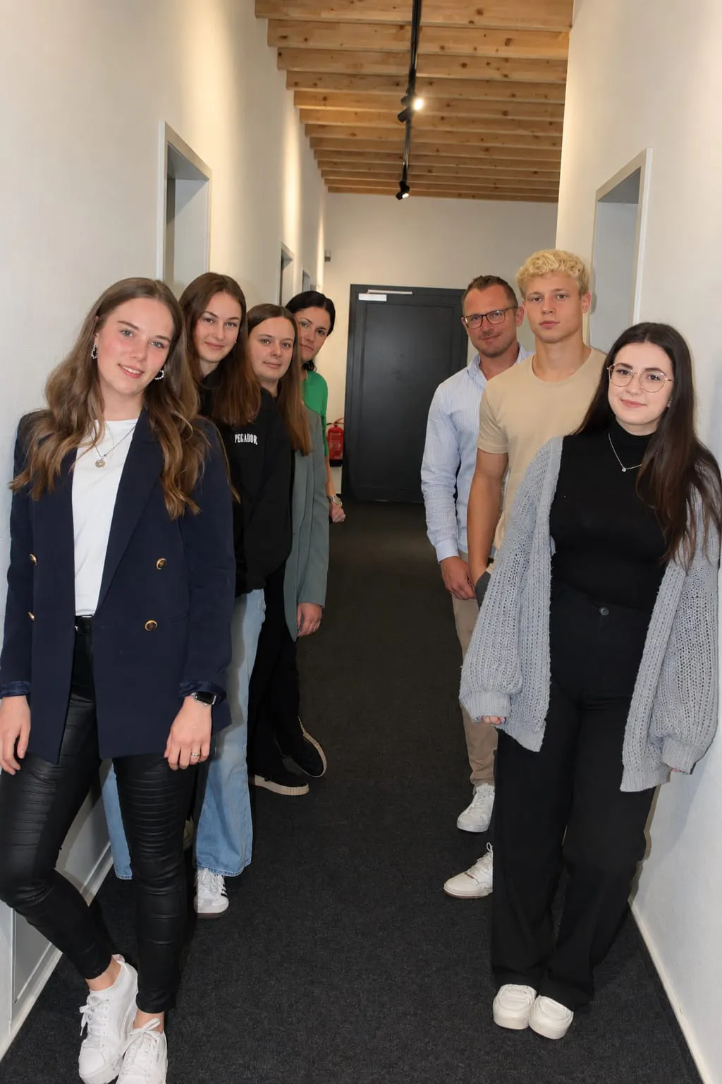 Sieben Personen stehen in einem Flur, drei Frauen links, vier Personen rechts, alle blicken zur Kamera, der Flur hat weiße Wände und eine Holzbalkendecke.