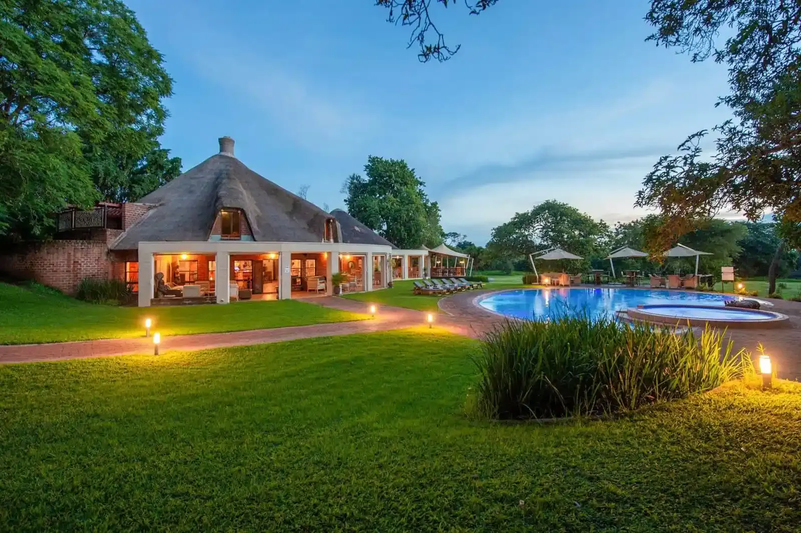 Thatched roof building with lit windows beside a round swimming pool surrounded by lawn, lounge chairs, umbrellas, and trees at dusk.