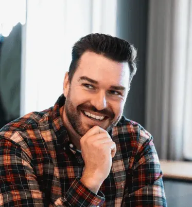 Smiling man with dark hair and beard wearing a red and black plaid shirt, resting his chin on his hand in a bright indoor setting.