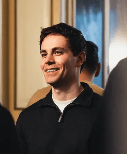 Young man with short dark hair wearing a black zip-up sweater, smiling and looking slightly to the left, with blurred background figures.