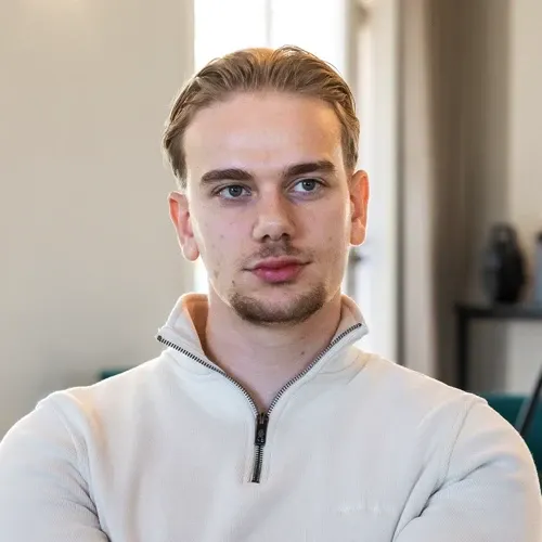 Young man with light brown hair wearing a white quarter-zip pullover, looking slightly to the side with a neutral expression in a softly lit indoor setting.