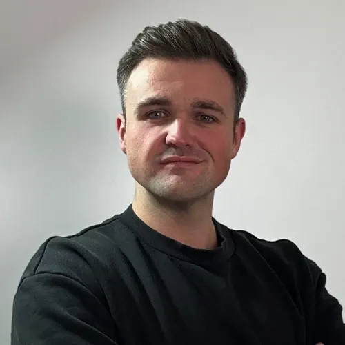 Man with short dark hair wearing a black shirt, facing forward with arms crossed against a plain gray background.