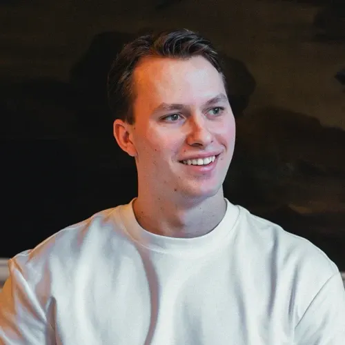 Young man with short brown hair wearing a white shirt, smiling and looking slightly to the right against a dark background.