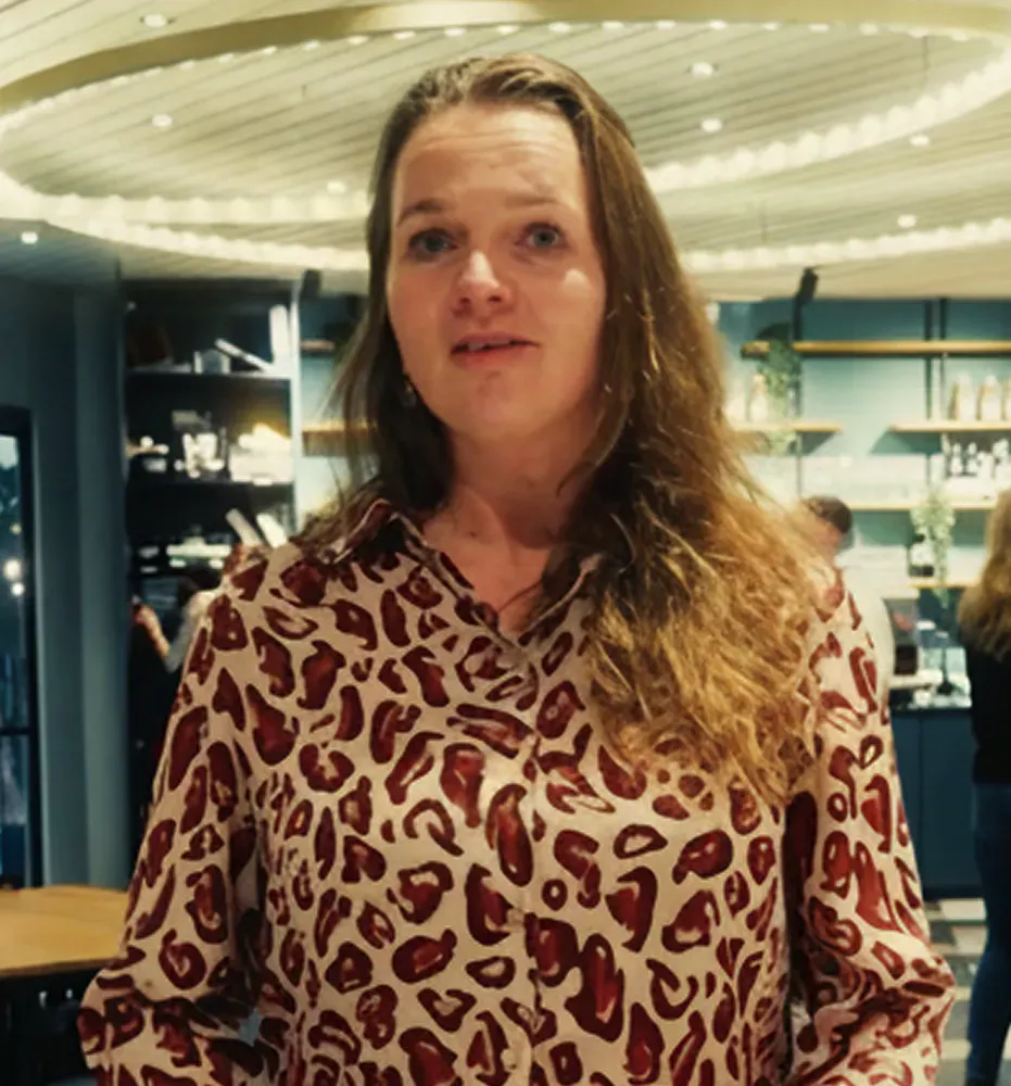 Woman with long wavy hair wearing a light-colored shirt with dark red leopard print, standing indoors with shelves and lights in the background.