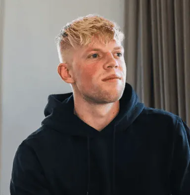 Young man with short blond hair wearing a black hoodie, looking slightly to the side, indoors with a neutral background and curtain.