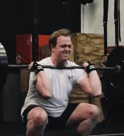 Man wearing gloves performing a front squat exercise with a barbell in a gym.