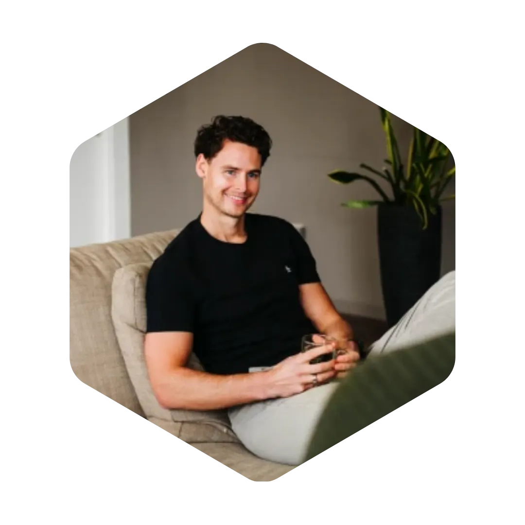 Young man with curly dark hair wearing a black t-shirt and light pants, sitting on a beige couch holding a glass, with a plant in a black pot in the background.