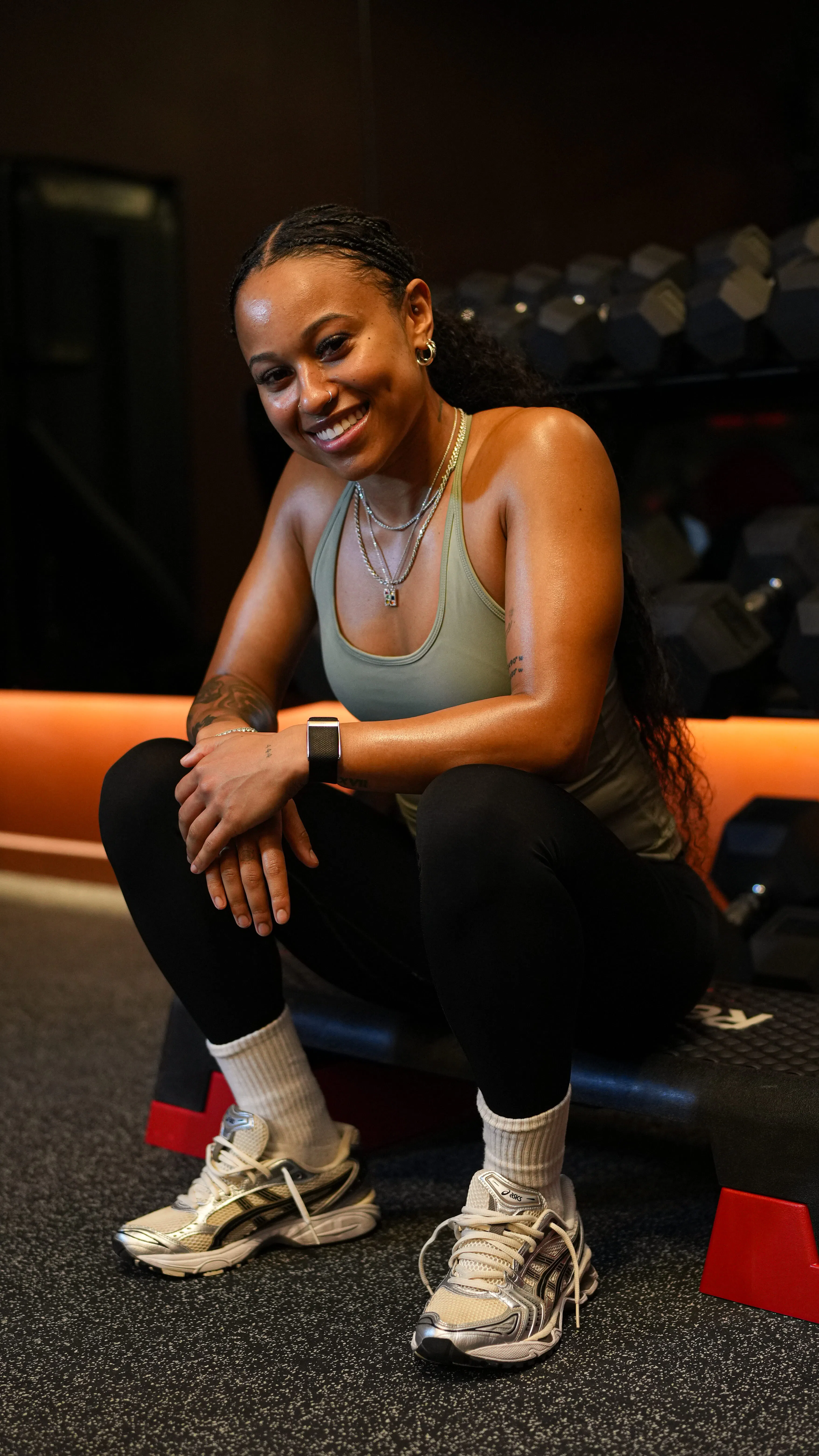 Smiling woman with braided hair in workout attire sitting on a step platform in a gym with dumbbells in the background.