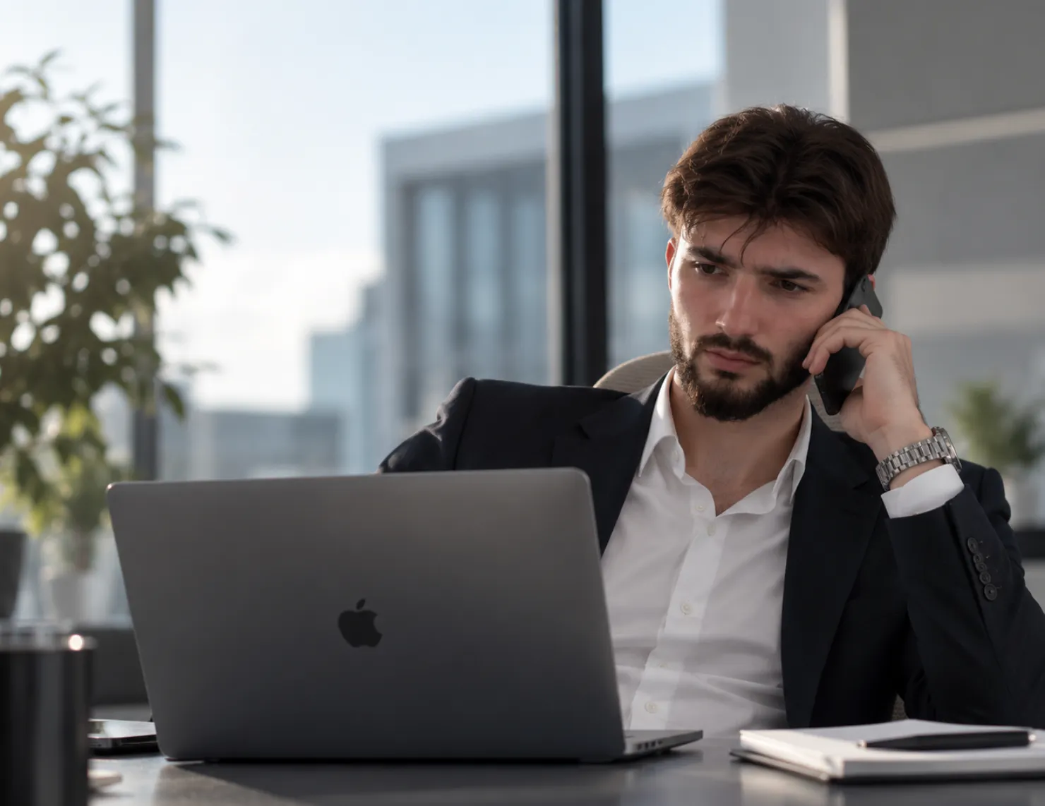 Mann in formeller Kleidung spricht am Telefon und arbeitet an einem Laptop in einem modernen Büro mit großen Fenstern und Tageslicht.
