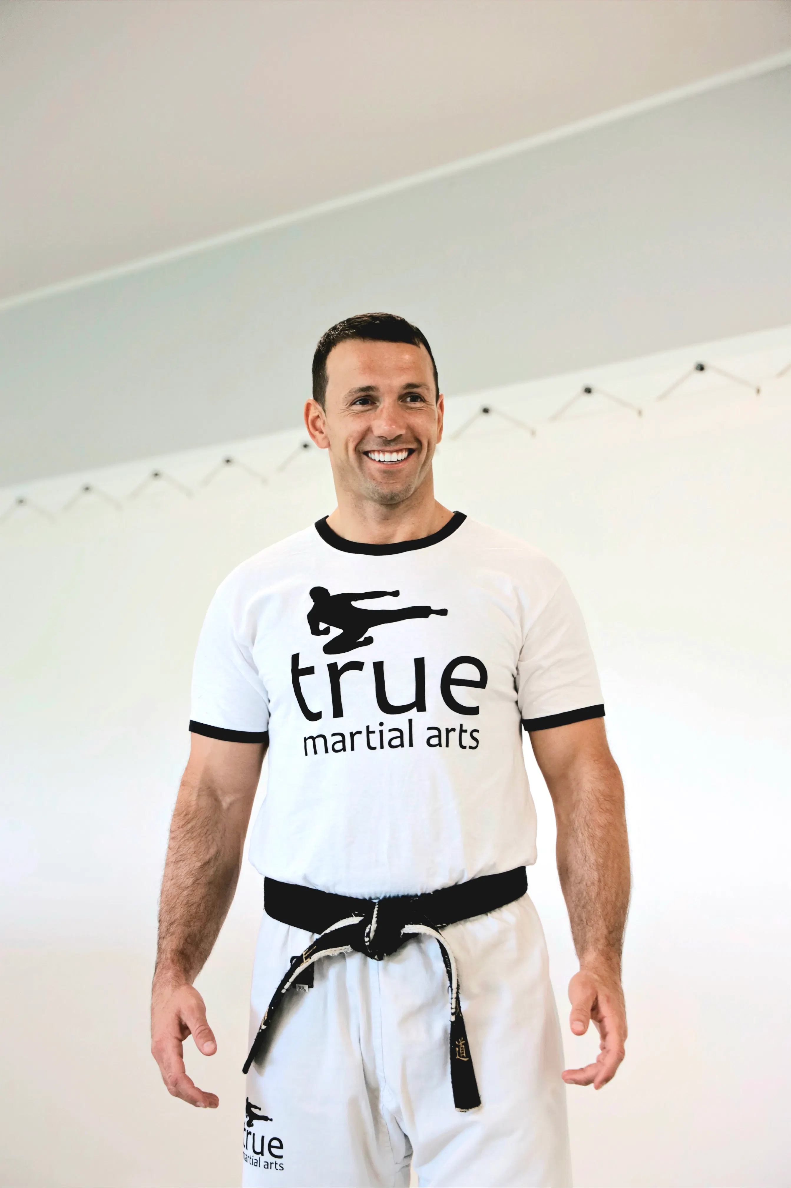 Mann in weißem T-Shirt und Hose mit schwarzem Gürtel lächelt, T-Shirt und Hose zeigen Aufdruck "true martial arts" mit Karate-Kämpfer-Grafik.