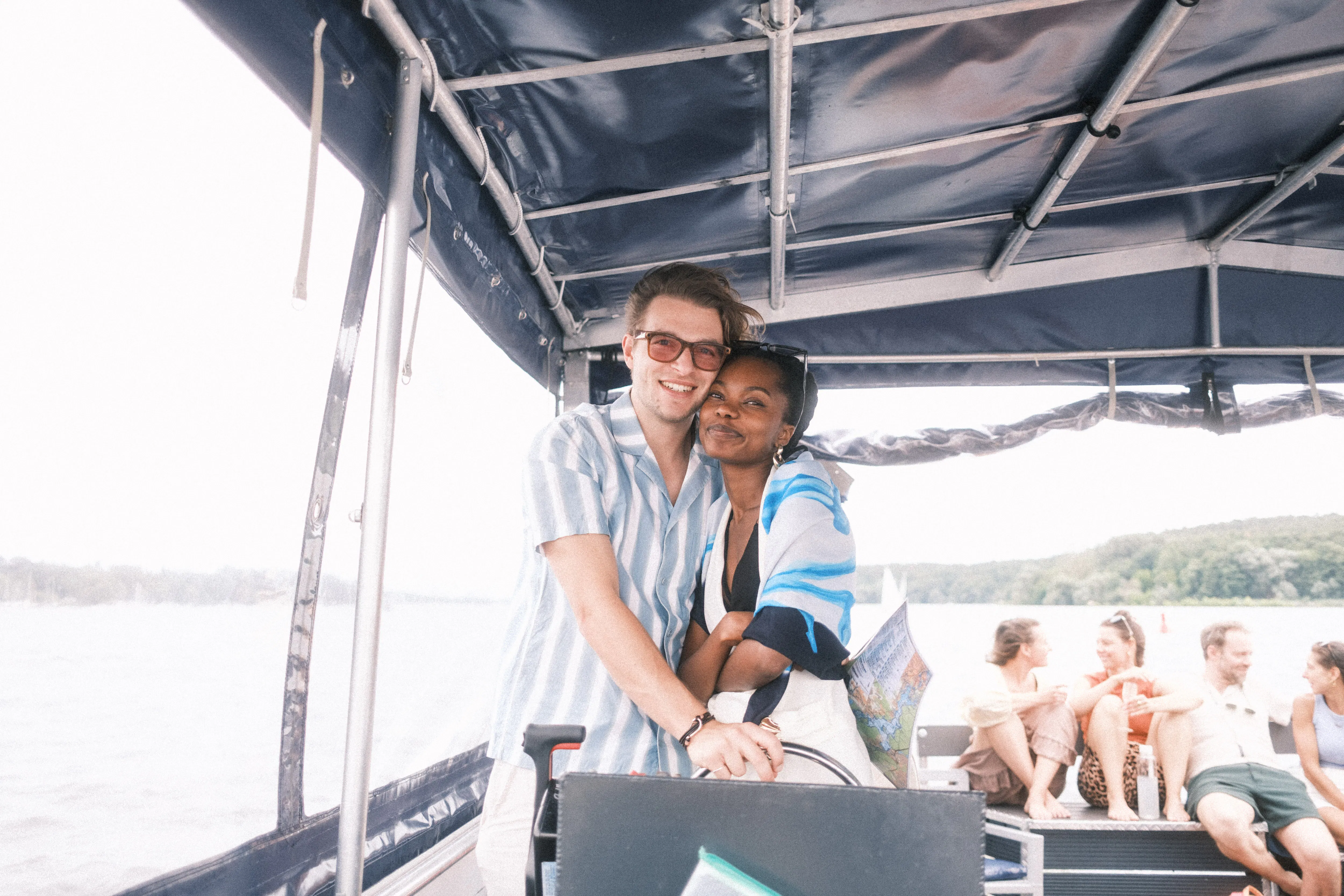 Two people smiling and embracing near boat controls under a canopy, with three others sitting and talking in the background on a boat on the water.