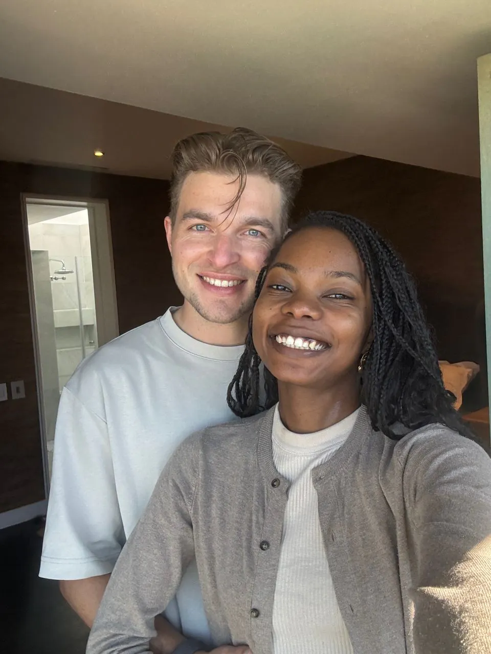 Smiling man and woman taking a selfie indoors, with the man standing behind the woman who is holding the camera.