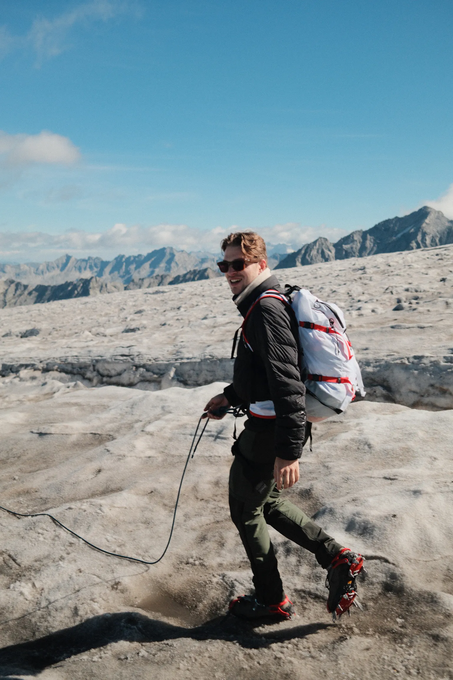 Man wearing hiking gear and crampons on icy terrain with mountains in the background under a blue sky.