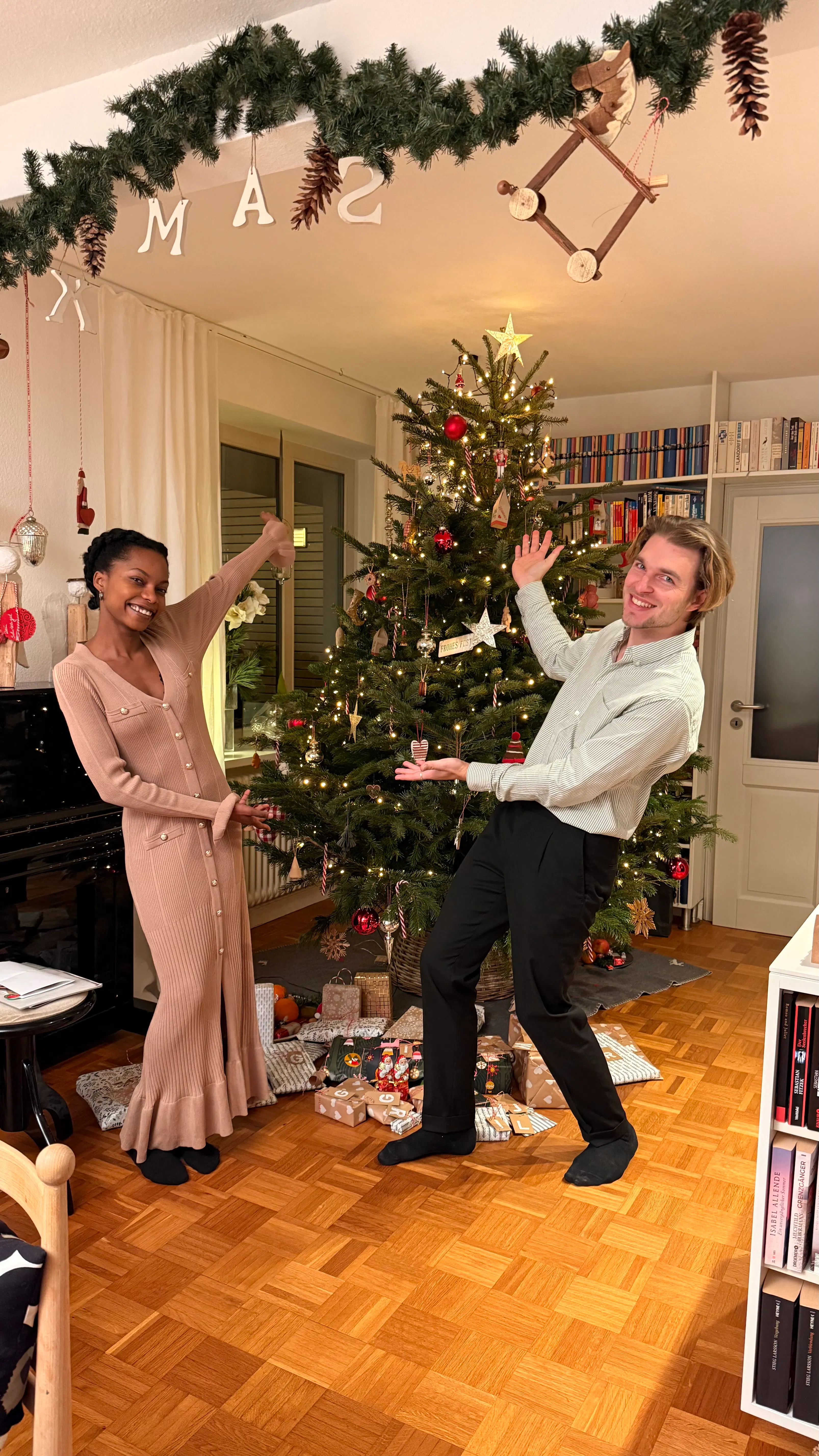 Two people smiling and gesturing towards a decorated Christmas tree with presents underneath in a cozy room with pine garland and wooden floor.