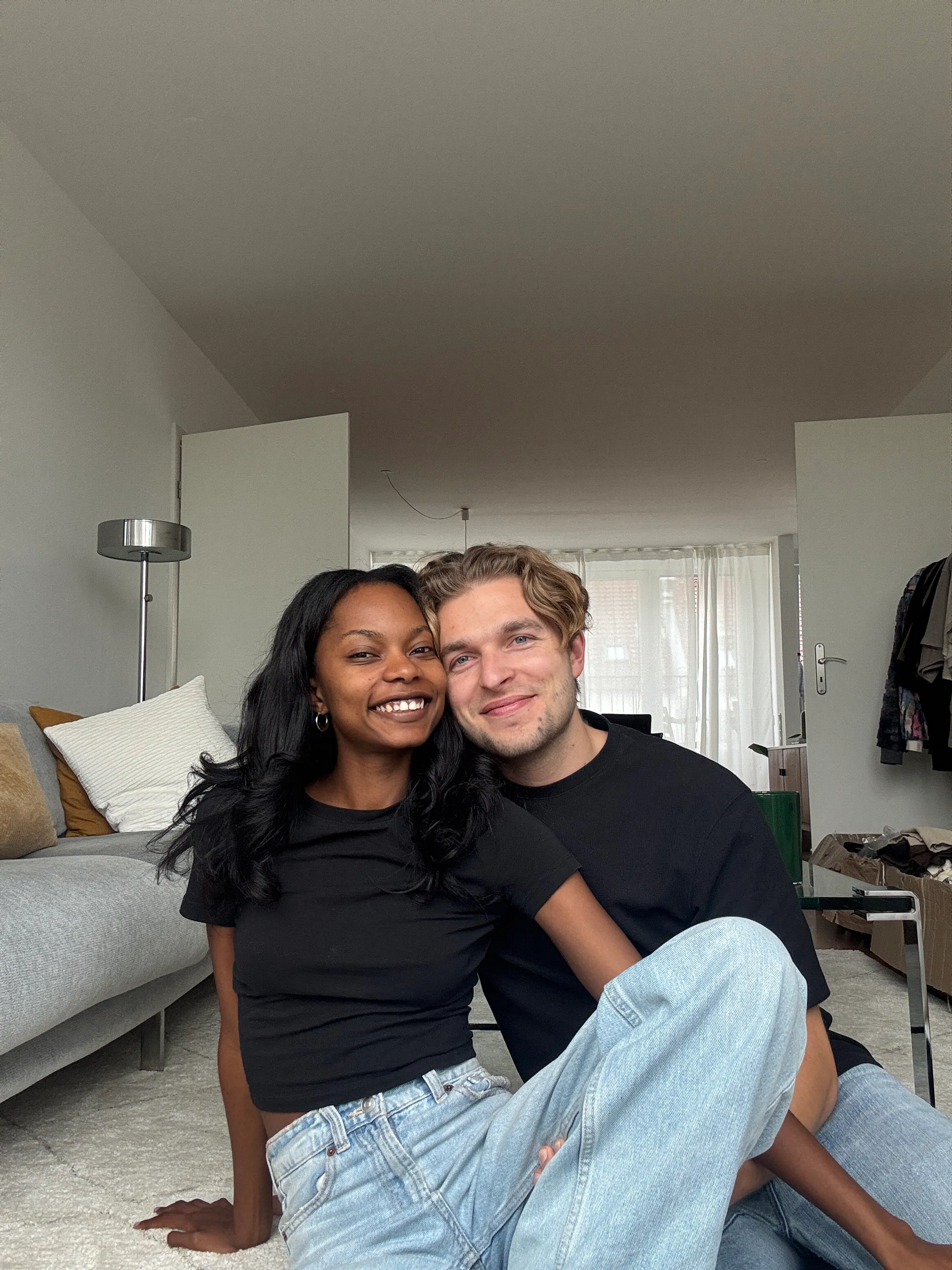 Smiling couple sitting on the floor in a living room, wearing casual black tops and jeans, with a sofa and open doors in the background.