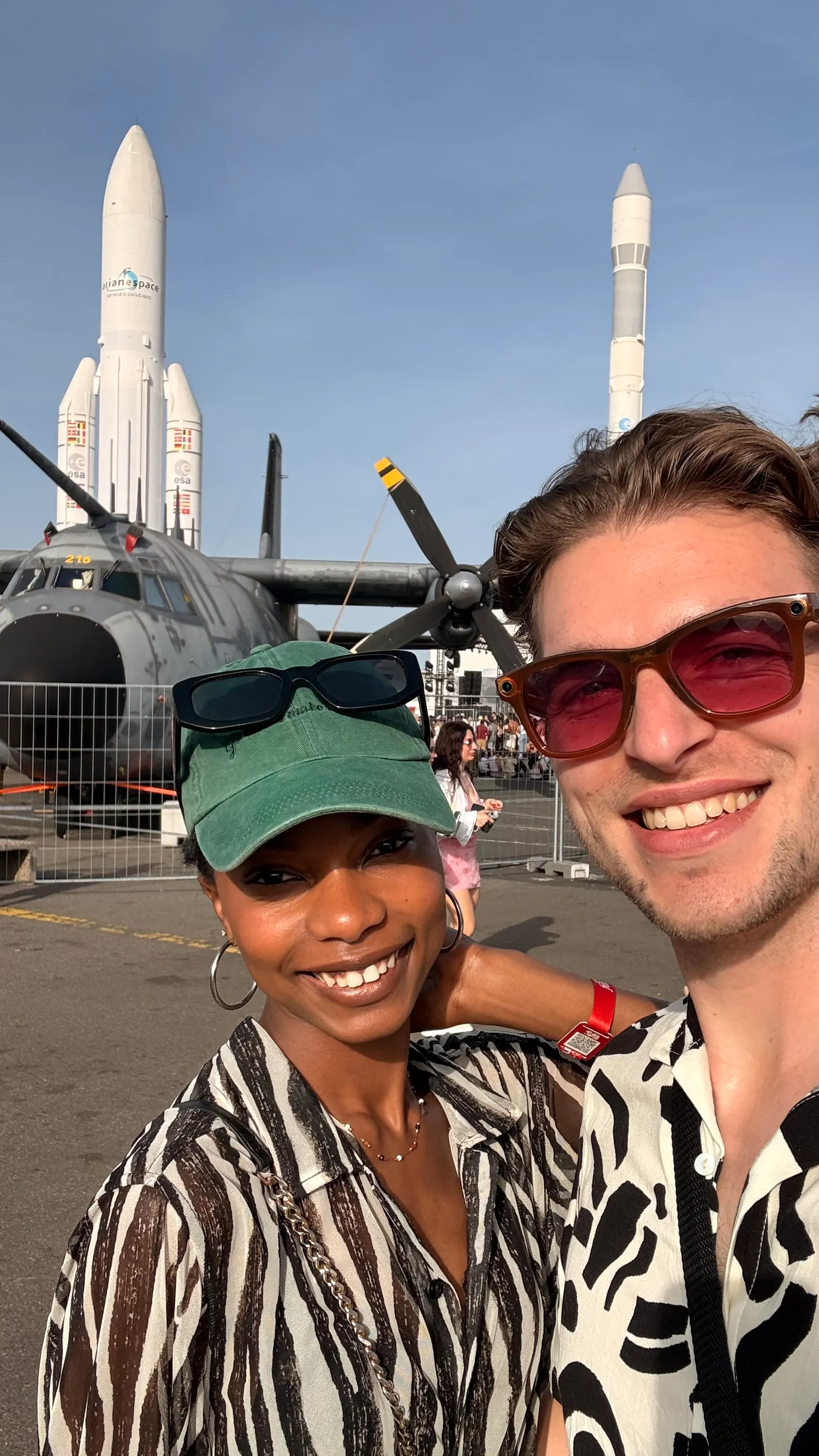 Two people smiling and taking a selfie outdoors with a propeller aircraft and two rockets in the background.