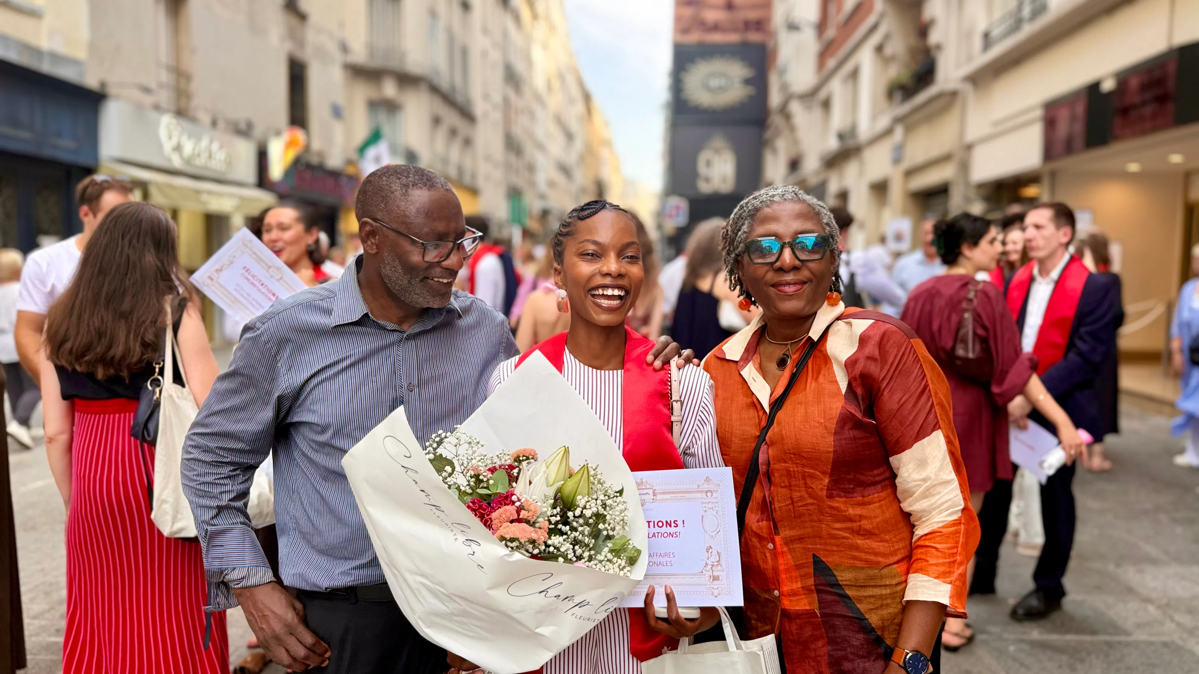 Young woman wearing a red graduation stole holds a bouquet and certificate, smiling between an older man in glasses and woman in colorful shirt on a city street with people in the background.
