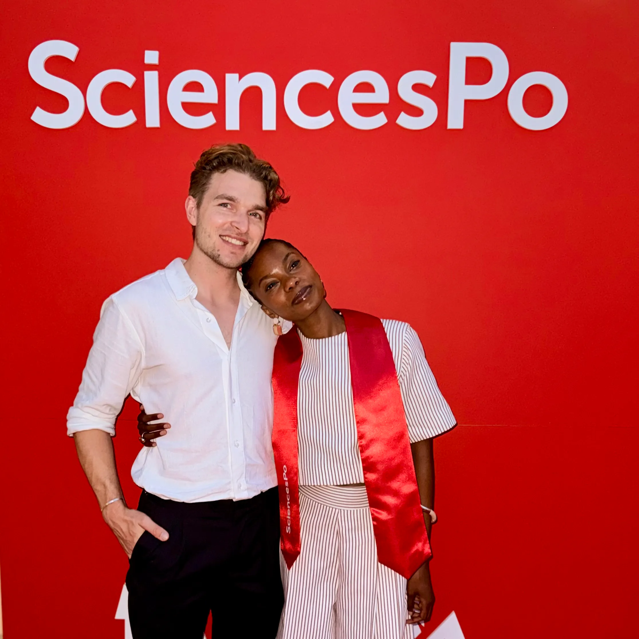 Two people smiling and posing in front of a red background with the word "SciencesPo," one wearing a white shirt and the other in a striped outfit with a red sash.