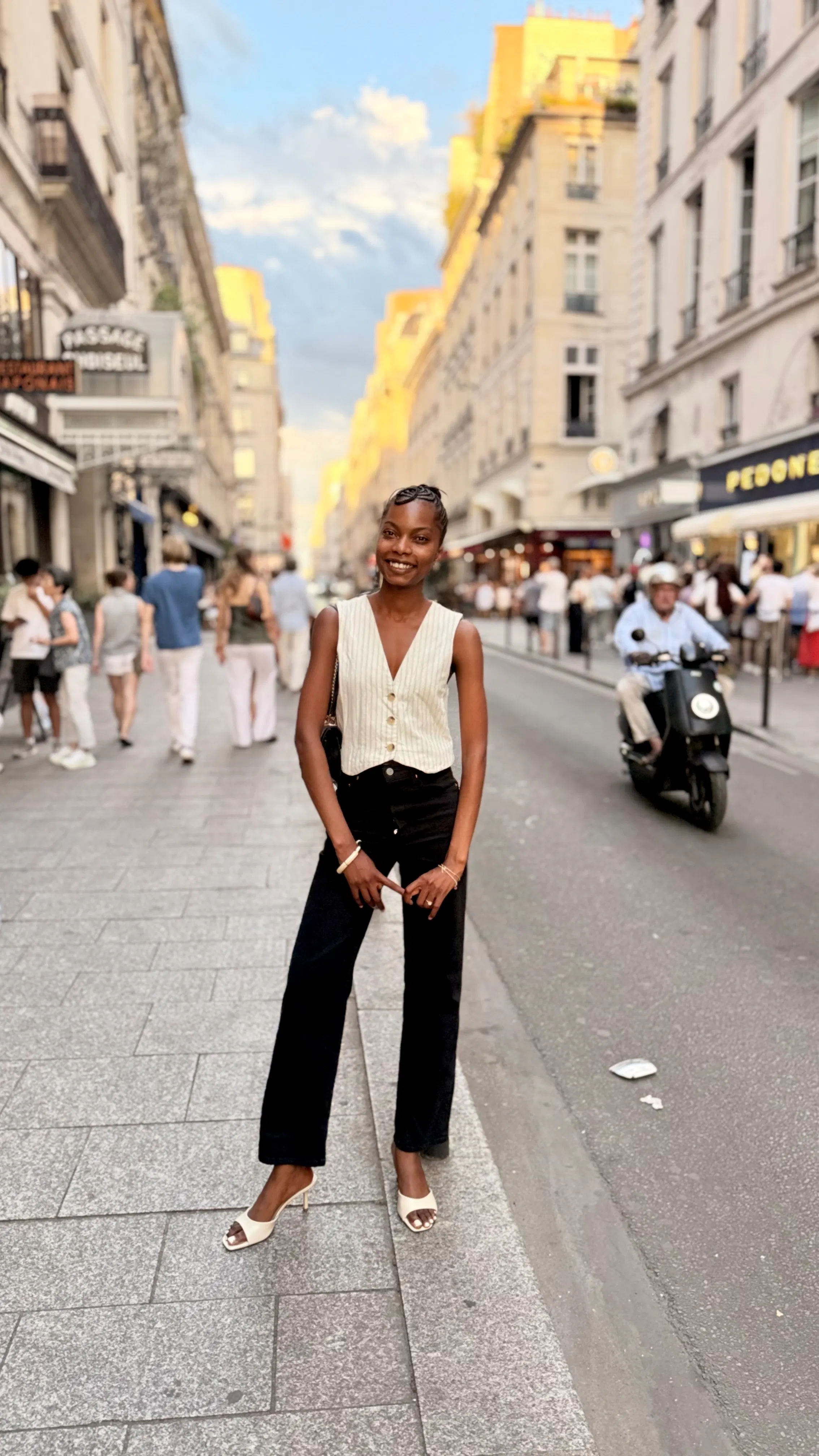 Young woman smiling and standing on a city street sidewalk wearing a sleeveless vest, black pants, and white heeled sandals, with people and buildings in the background.