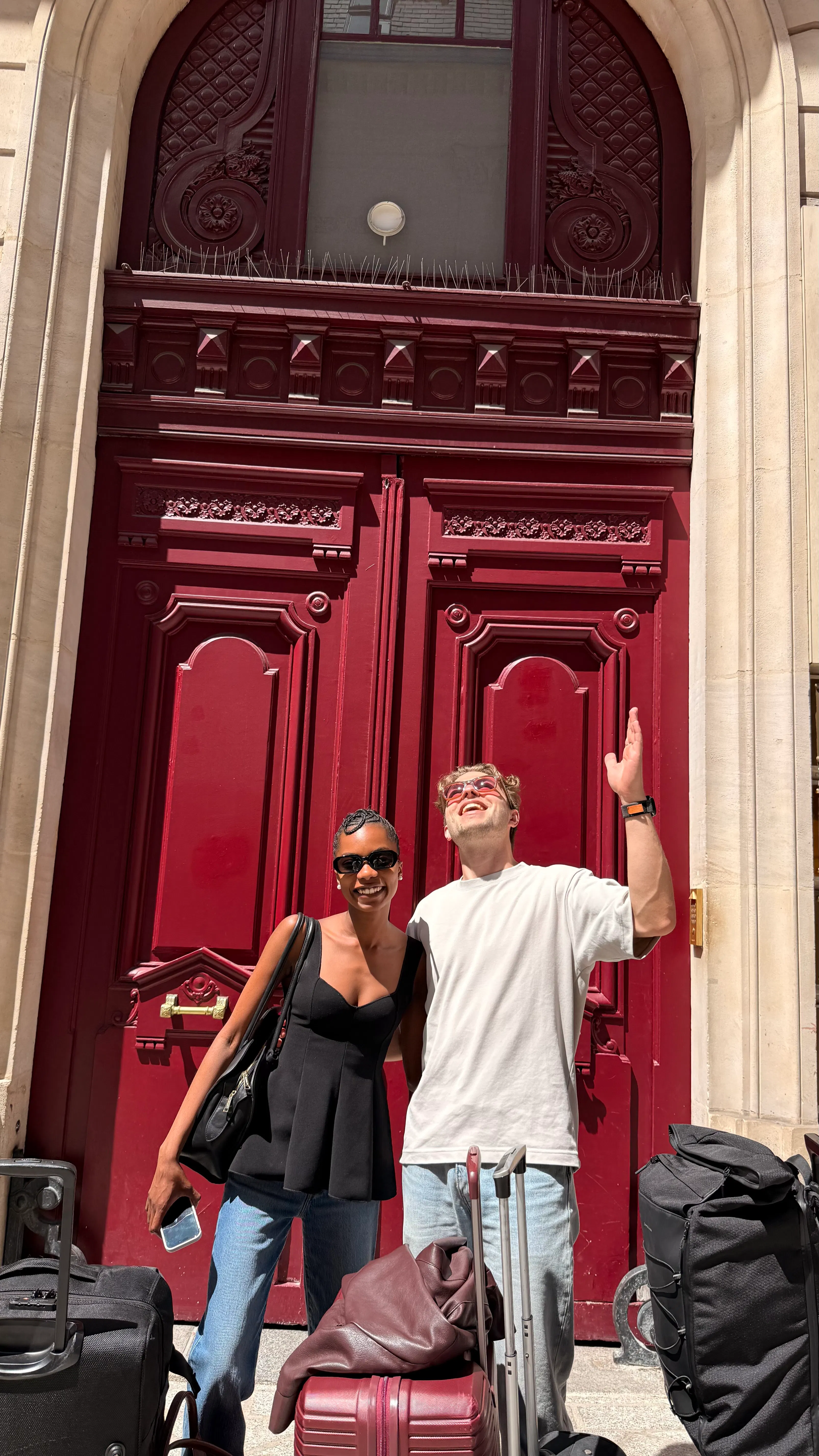 A smiling man and woman wearing sunglasses stand with suitcases in front of a large ornate red door, the man gestures upwards while the woman holds a smartphone.