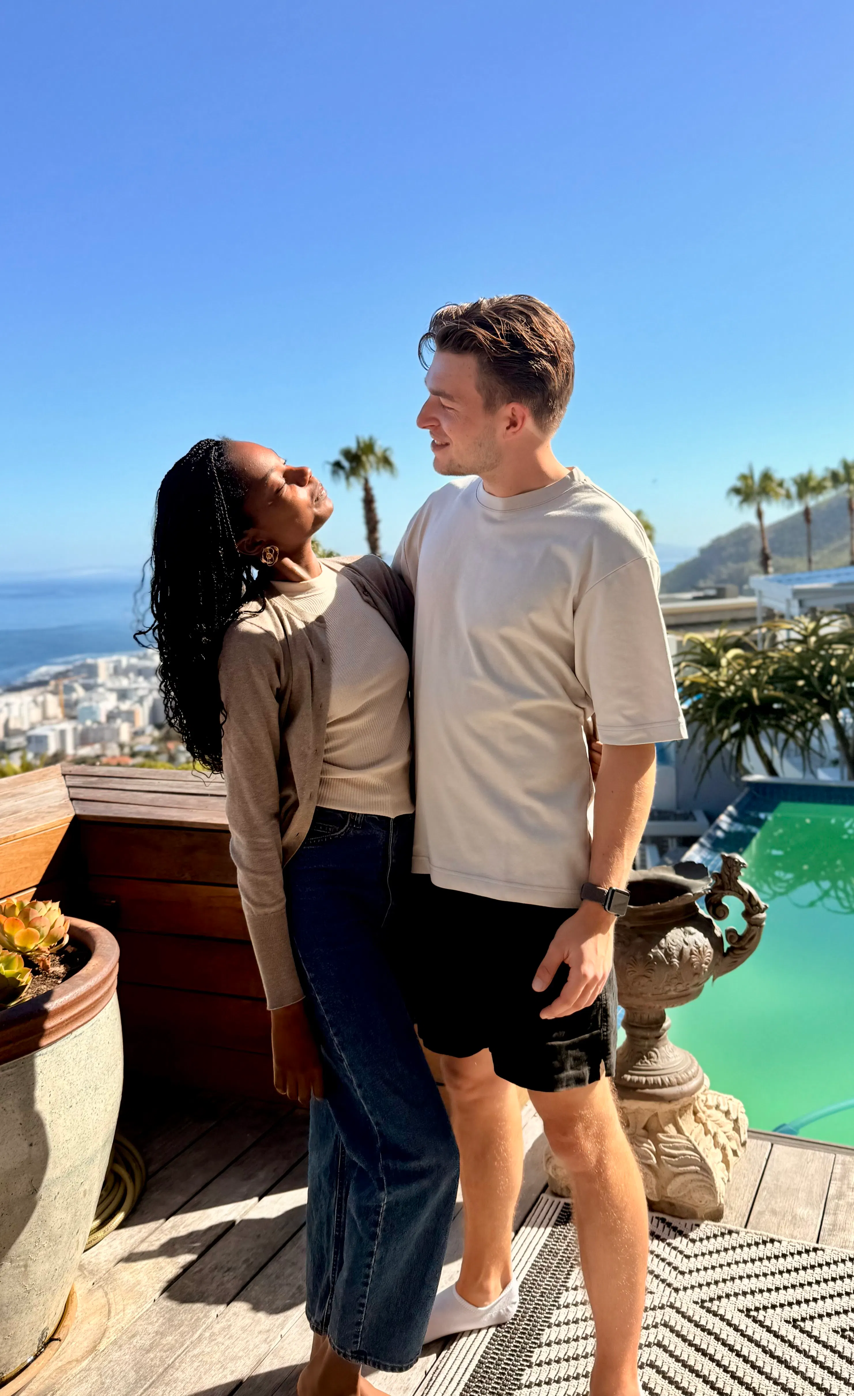 Couple standing on a wooden deck by a pool, looking at each other with a city, ocean, and palm trees in the background under a clear blue sky.