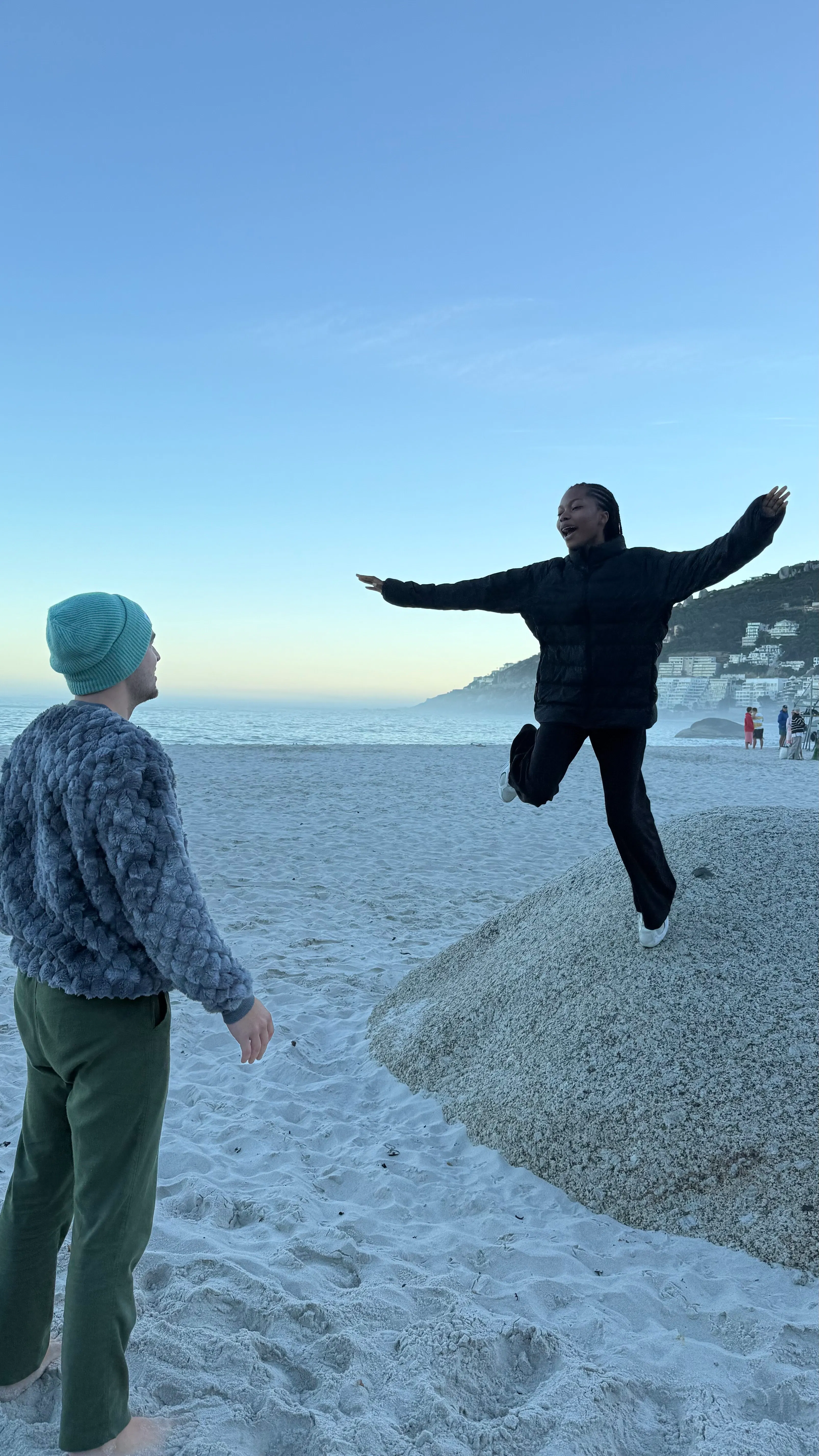 Person wearing a black jacket standing with one leg raised on a large rock on the beach, facing another person in green pants and a blue beanie, with ocean and hills in the background.
