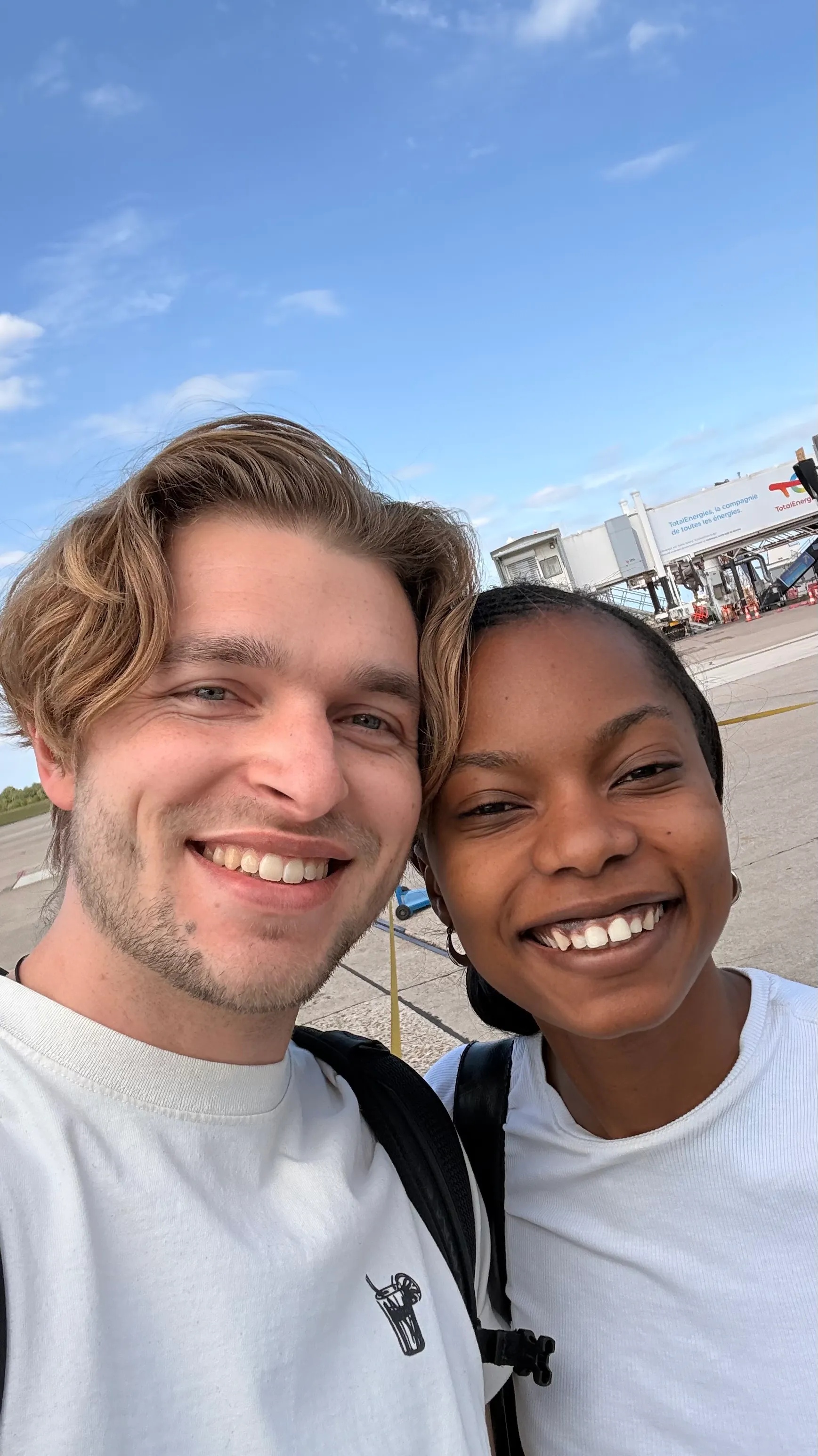 Two smiling people wearing white shirts and backpacks taking a selfie at an airport tarmac with terminal gates and blue sky in the background.