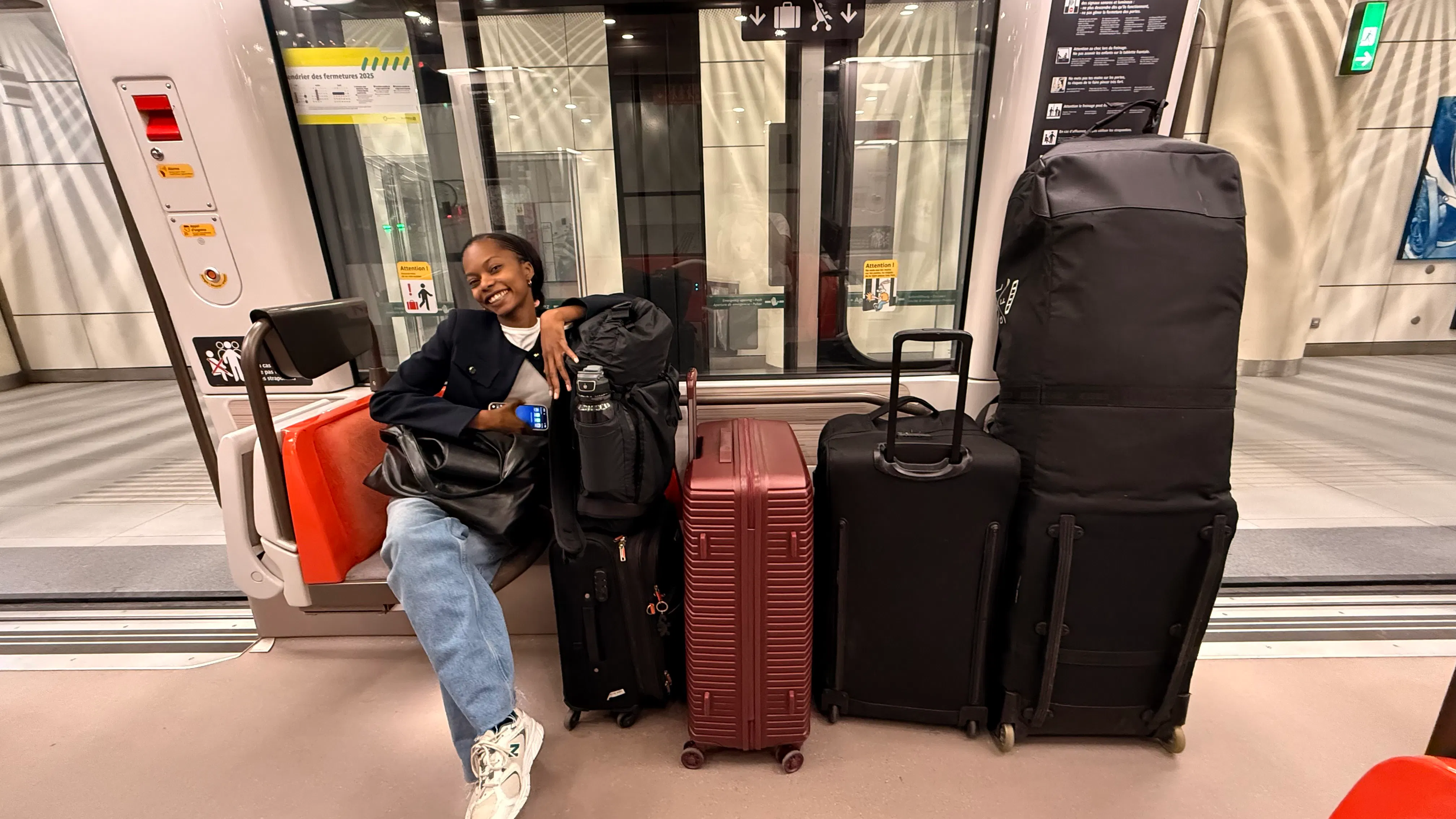 Young woman smiling and seated in a subway car, surrounded by four large suitcases and bags, with subway platform visible through glass doors behind her.