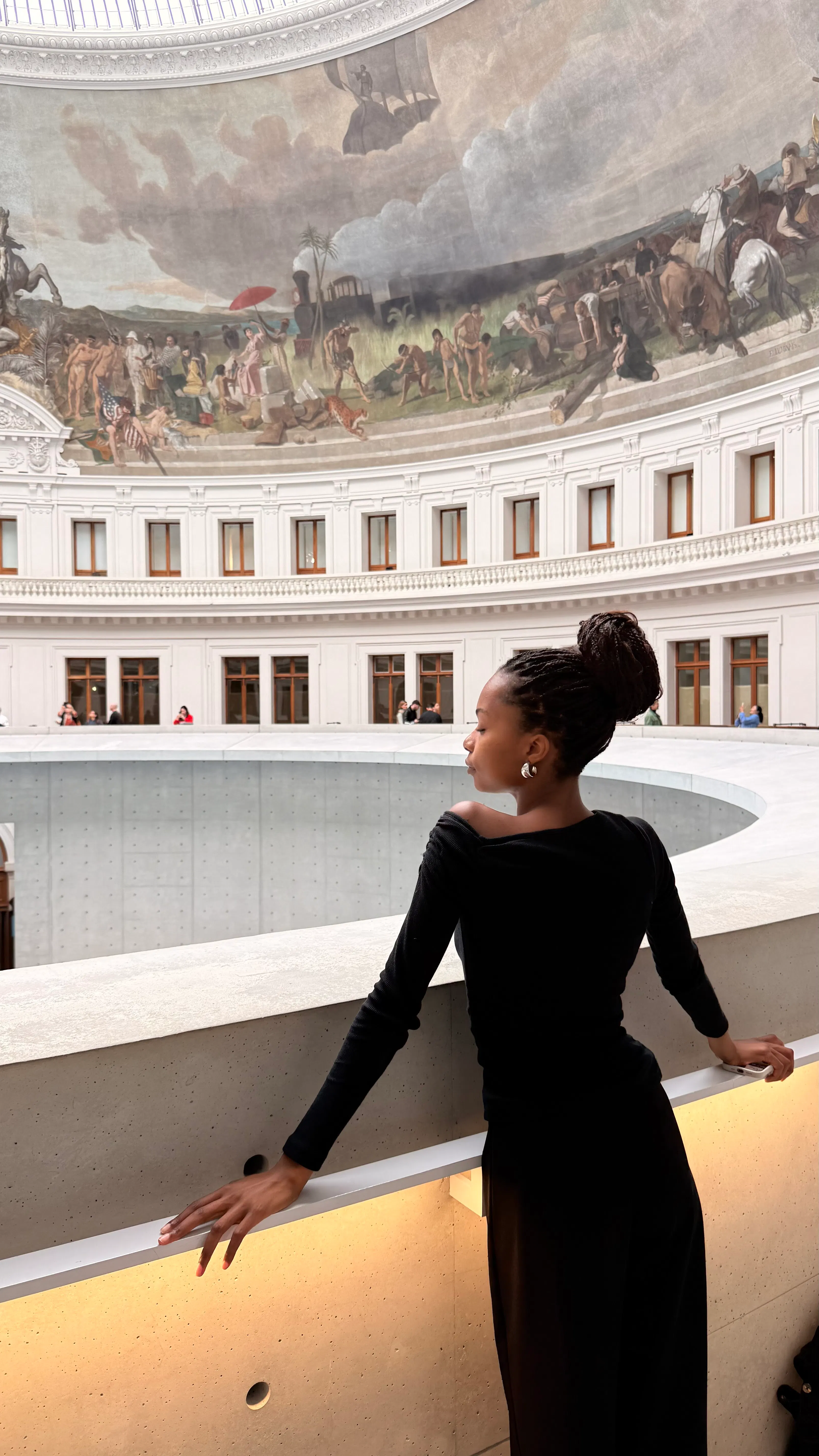 Woman in black outfit with braided hair in a bun leaning on a railing inside a circular gallery with large windows and a mural depicting historical scenes above.