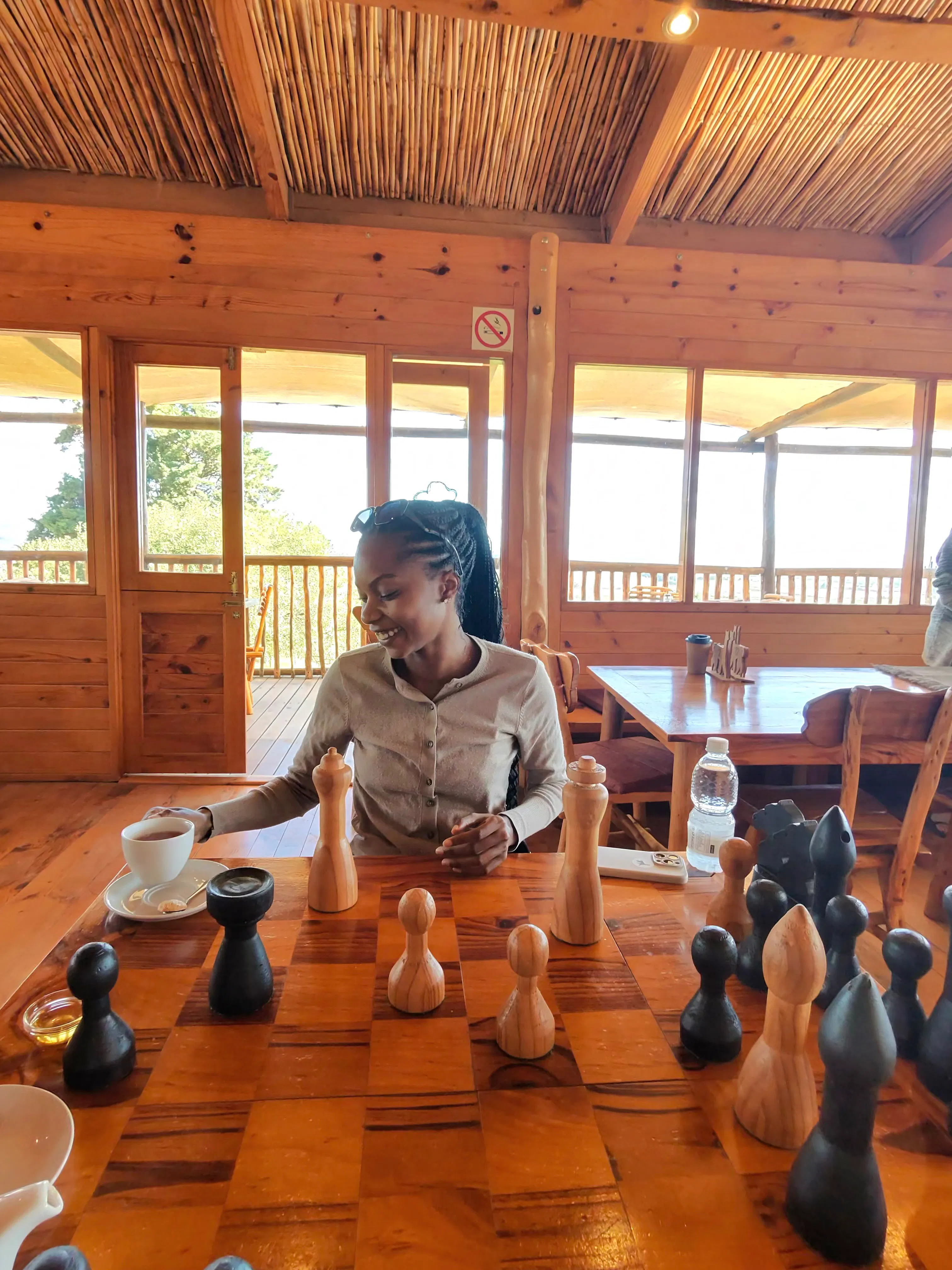 Woman smiling and reaching for a cup while sitting at a large wooden chessboard with oversized black and wooden chess pieces in a wood-paneled room with large windows.