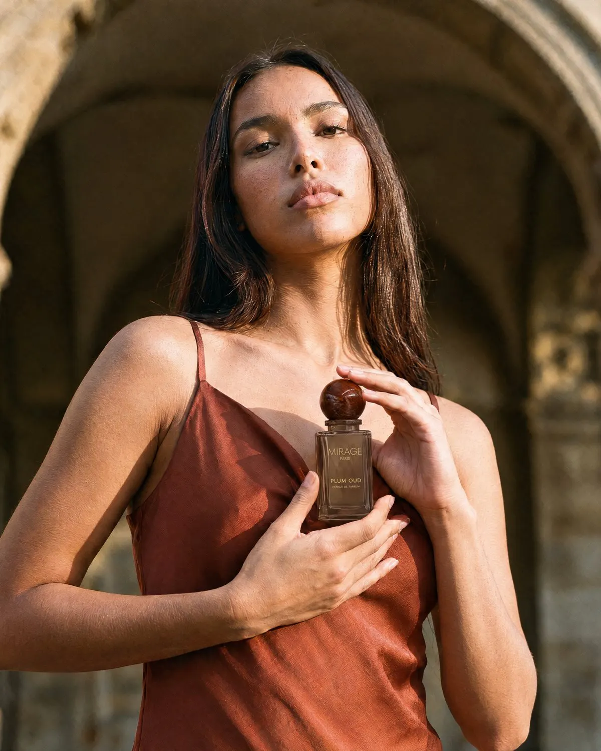 Frau in einem rostfarbenen ärmellosen Kleid hält eine braune Parfümflasche mit der Aufschrift "Mirage Paris Plum Oud" vor einer steinernen Bogenstruktur.