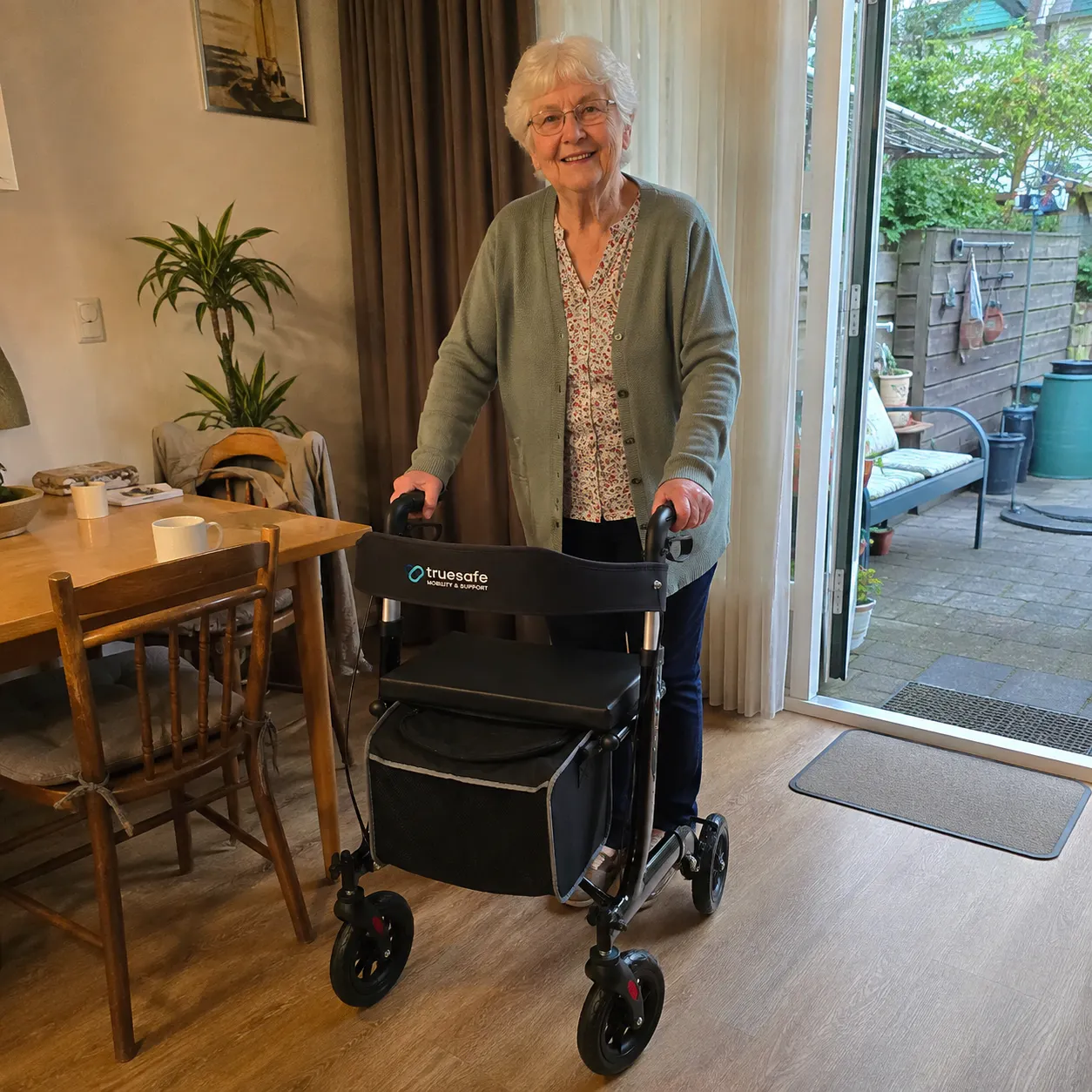 Elderly woman indoors using a black truesafe mobility walker with four wheels and a storage bag, standing near a dining table and an open door to a garden.