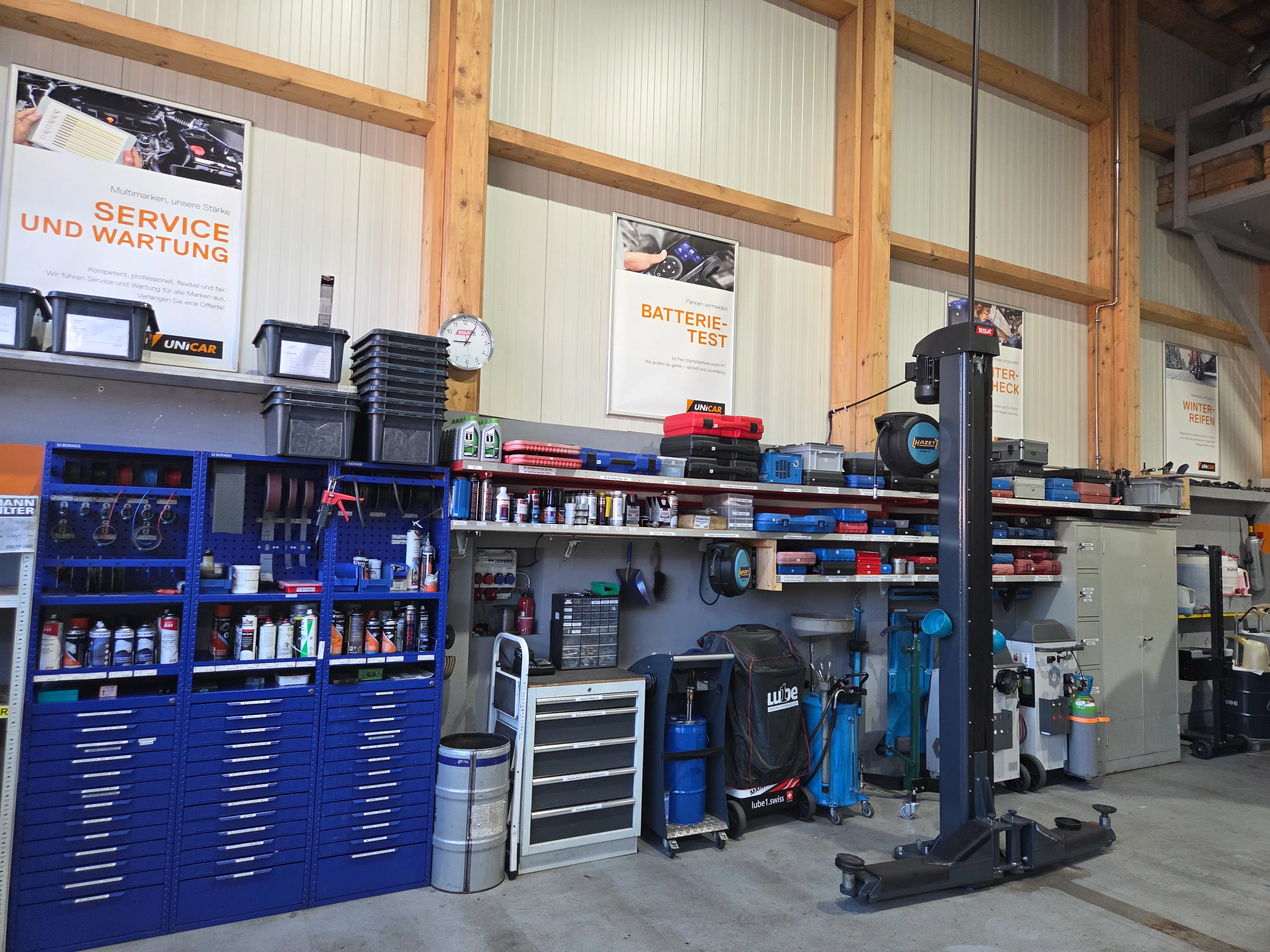 Werkstatt mit organisiertem Werkzeugschrank, Regalen mit Dosen und Werkzeugkisten, Autolift und Wandschildern zu Service, Batterie-Test, Batteriezustand und Winterreifen.