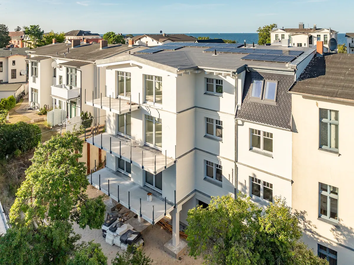 Mehrstöckiges Wohngebäude mit Balkonen unter sonnigem Himmel, einige Bauarbeiten sichtbar, Solarpaneele auf dem Dach, Bäume und andere Gebäude in der Nähe, Meer im Hintergrund.